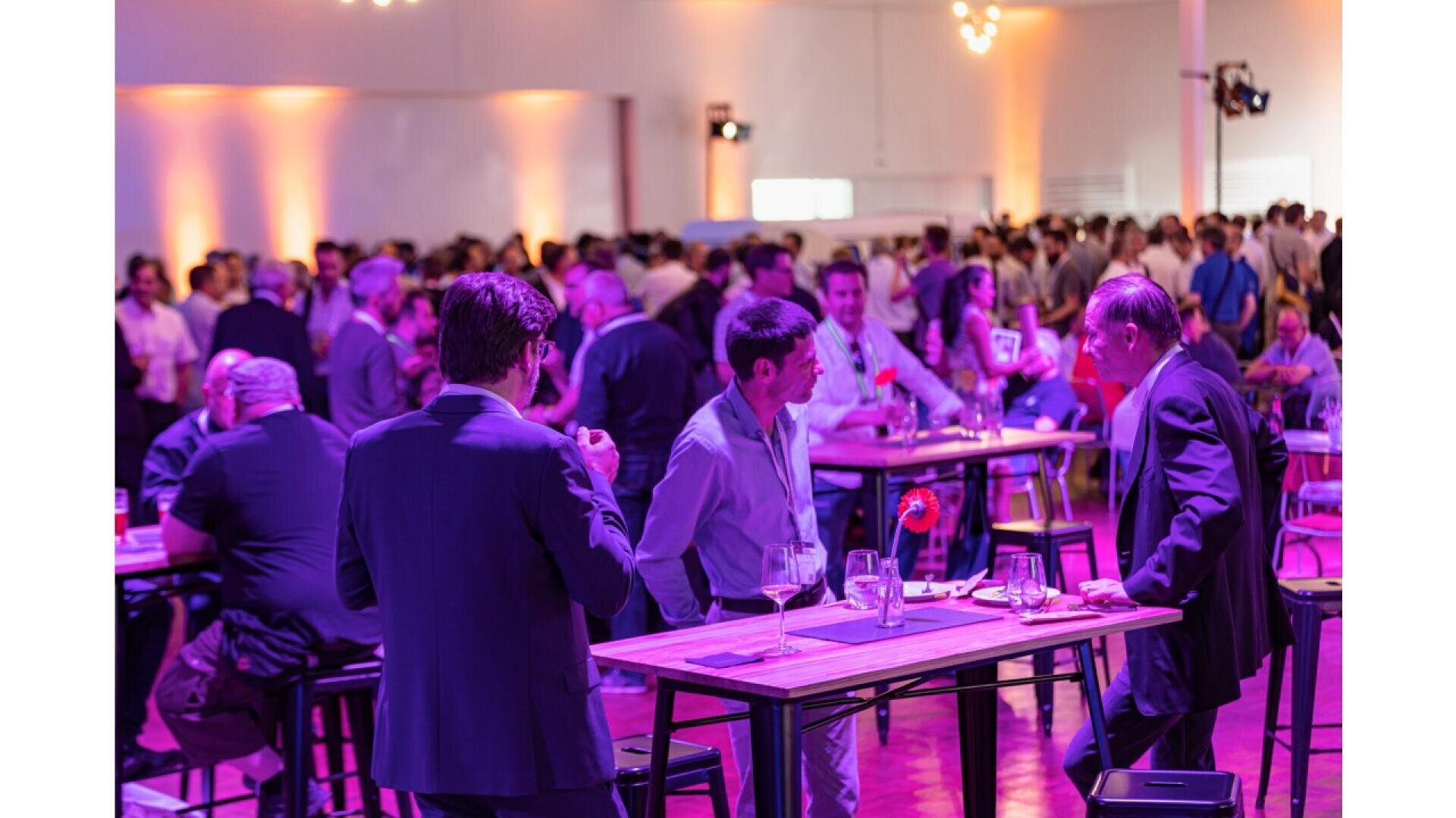 A large group of people mingling at a lively indoor event, gathered around tall tables with drinks in hand. The room is lit with purple and orange lights, creating a festive and energetic atmosphere.