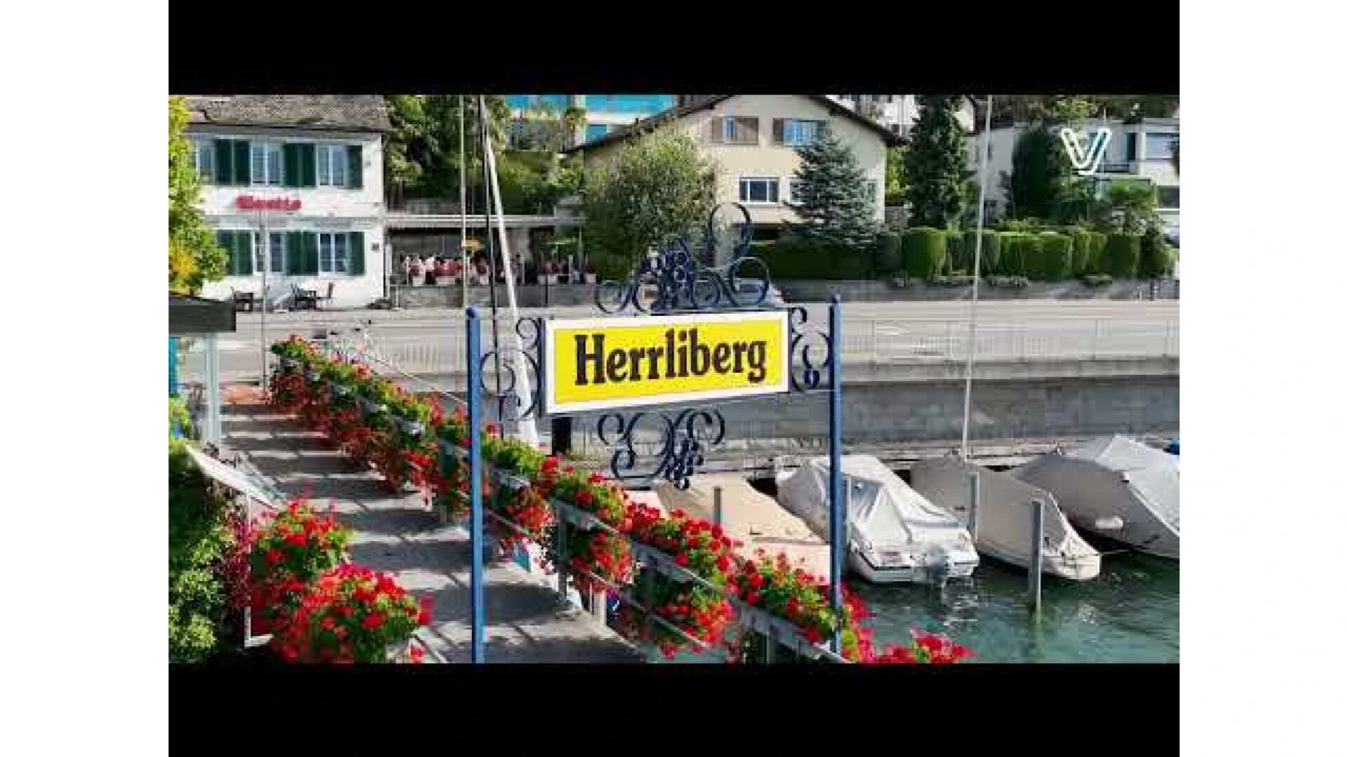 Ein blau-gelbes Schild mit der Aufschrift Herrliberg steht auf einem mit roten Blumen gesäumten Steg, mit Booten auf dem Wasser und Häusern im Hintergrund.