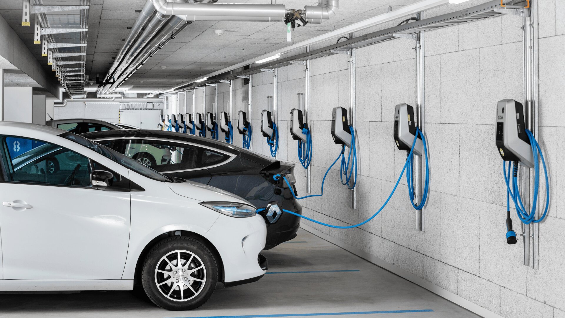 In einer modernen Tiefgarage sind mehrere Elektroautos nebeneinander geparkt, die mit blauen Kabeln an Ladestationen an der Wand angeschlossen sind. Die Umgebung wirkt sauber und gut beleuchtet.