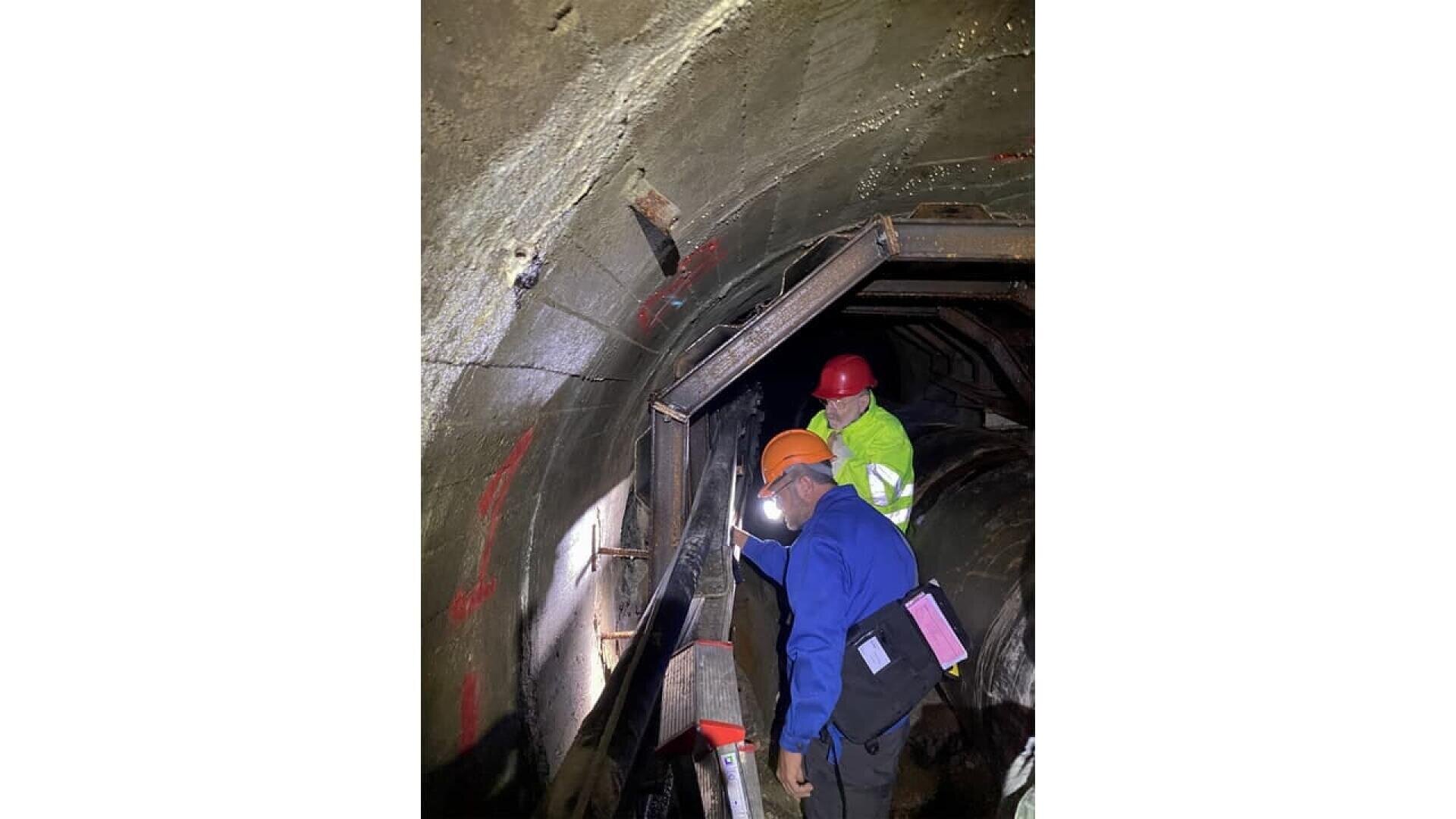 Two workers wearing safety gear and helmets inspect the inside of a dimly lit tunnel, examining the wall and using flashlights. Red markings are visible on the concrete surface.