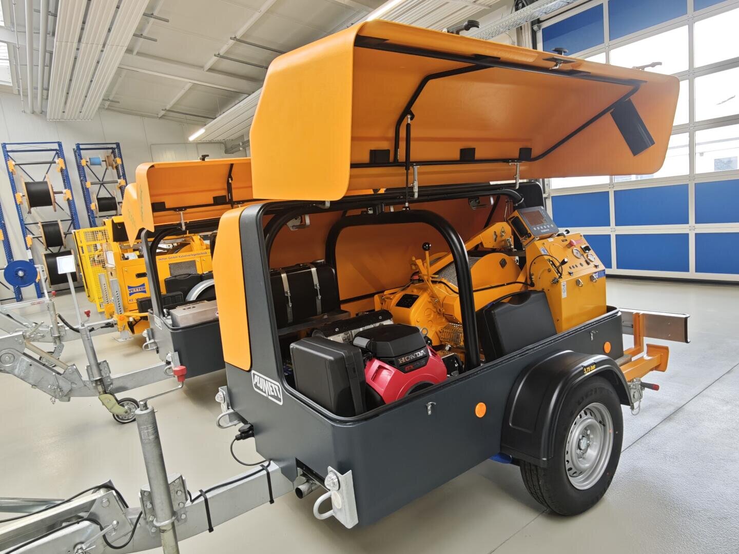 In a clean, well-lit workshop with blue and white walls and large windows stand two mobile industrial machines, their orange covers open to reveal the mechanical components inside.