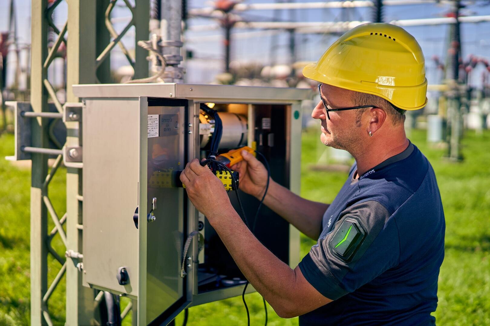 Ein Mann, der einen gelben Schutzhelm und eine Schutzbrille trägt, arbeitet an einem sonnigen Tag mit Werkzeugen an elektrischen Geräten in einem Metallschaltkasten in einem Kraftwerk.