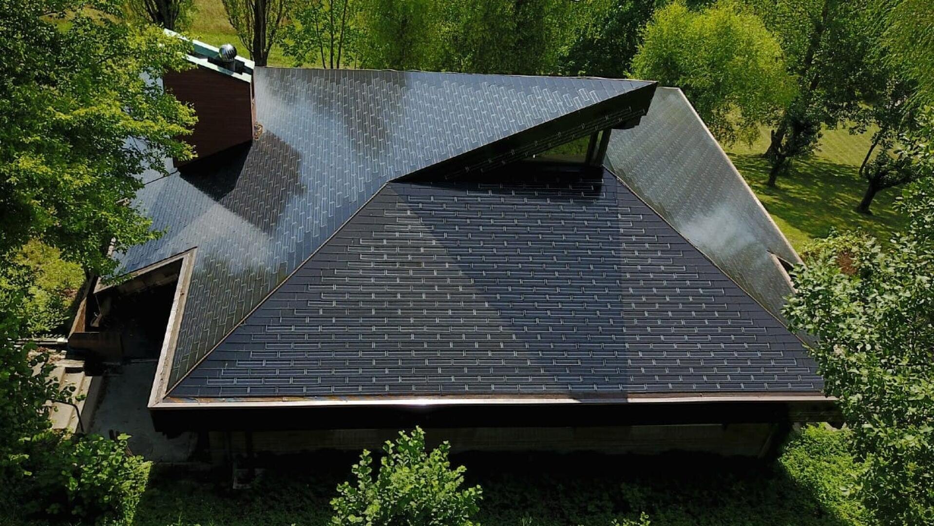 Ein mit dunklen, glänzenden Solarschindeln gedecktes Hausdach, umgeben von grünen Bäumen und Gras, von oben betrachtet an einem sonnigen Tag.