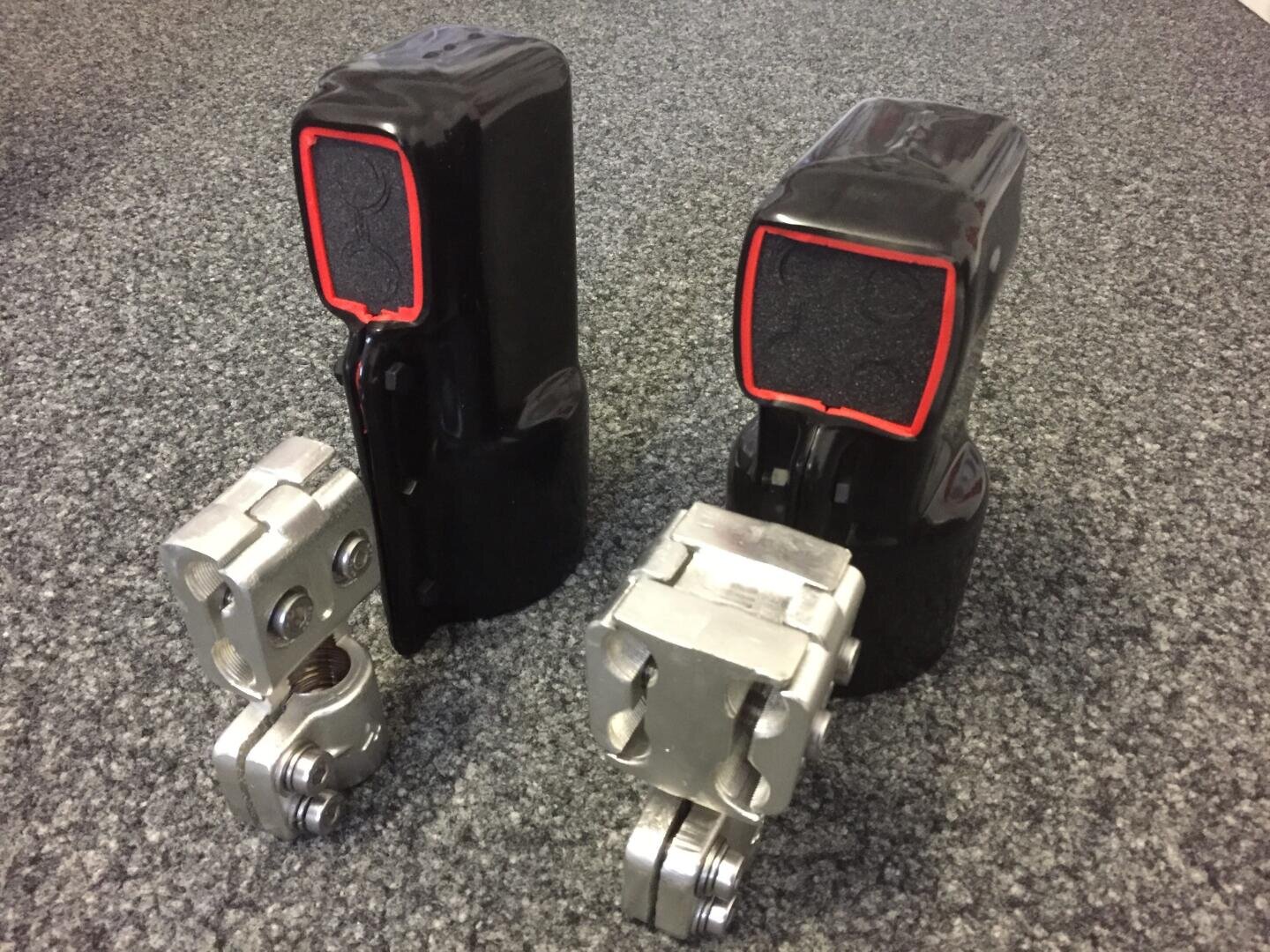 Zwei mechanische Metallgelenke befinden sich vor zwei schwarzen, rechteckigen Geräten mit rot umrandeten Frontplatten, die auf einem grau gesprenkelten Teppich stehen.