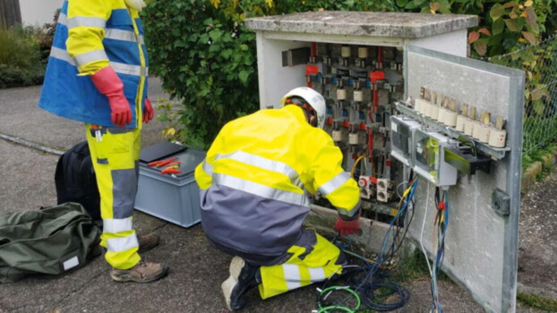 Two utility workers in high-visibility gear and helmets work on an open electrical or communications box outdoors, surrounded by tools, wires, and equipment. One kneels while the other stands nearby.