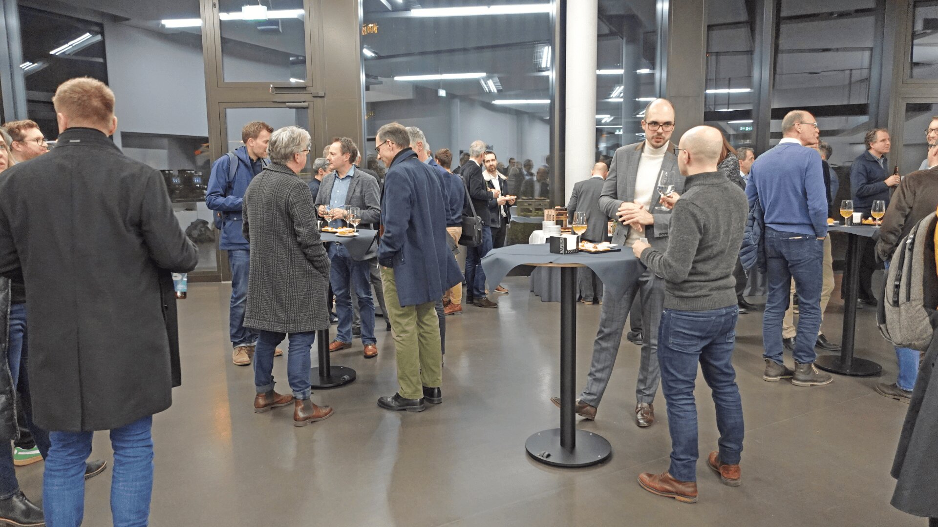 Eine Gruppe von Geschäftsleuten sitzt an hohen Tischen mit Getränken und Snacks in einem modernen, hellen Innenraum mit großen Fenstern und knüpft Kontakte.