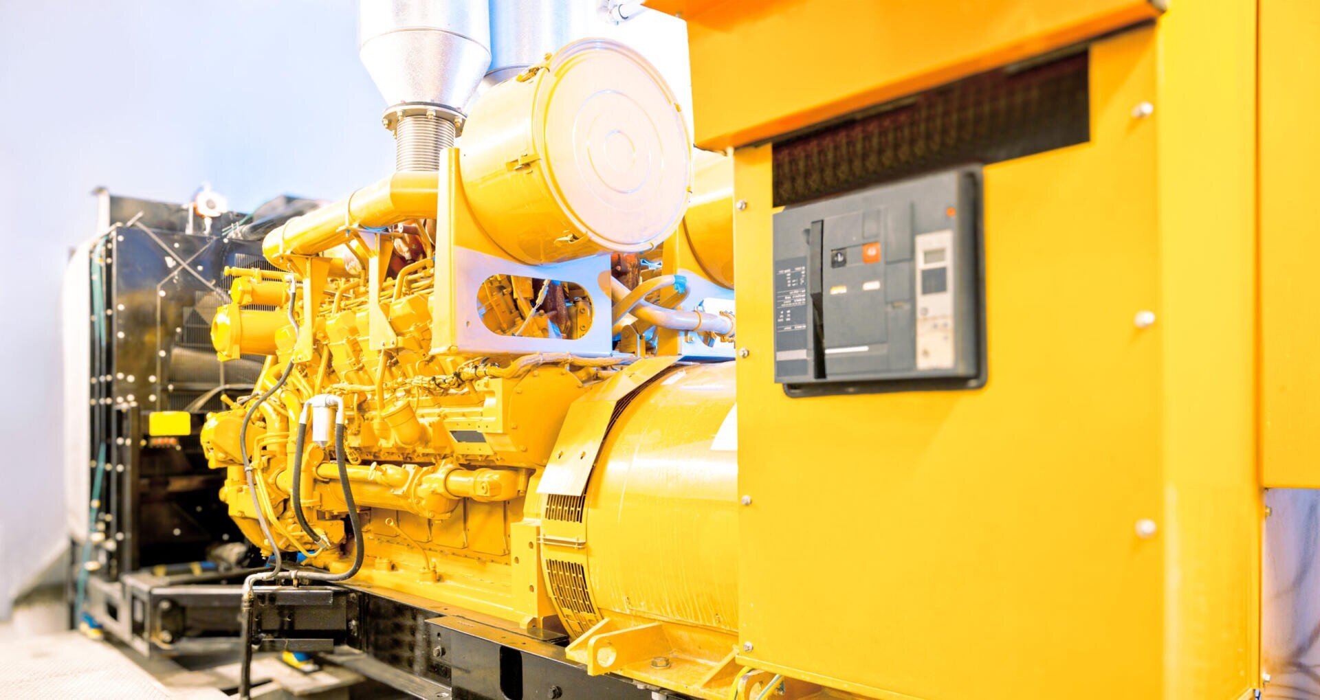 A large yellow generator with intricate yellow pipes and a silver exhaust pipe, featuring a black control panel with a grey circuit breaker.