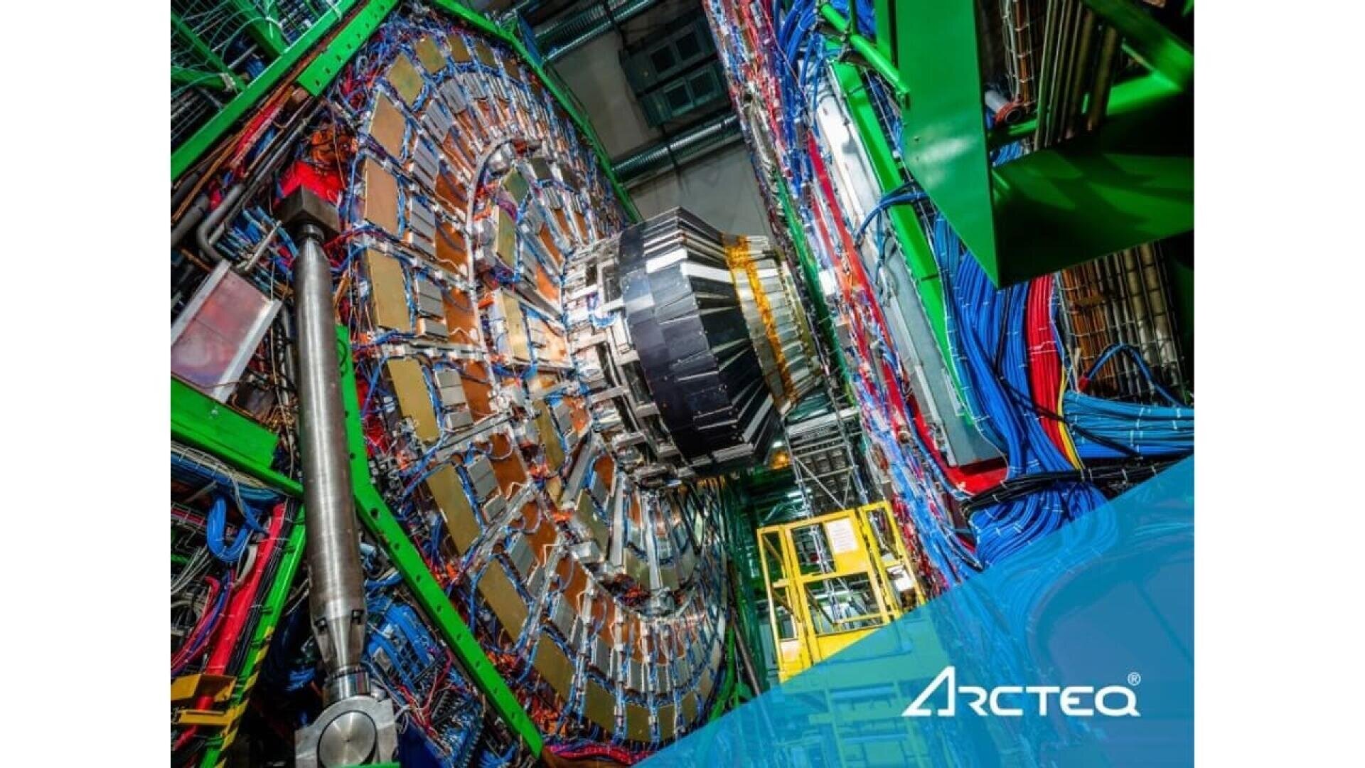 A large, circular particle detector filled with colorful cables and metal parts inside a laboratory. The foreground features the ARCTEQ logo with blue geometric shapes.