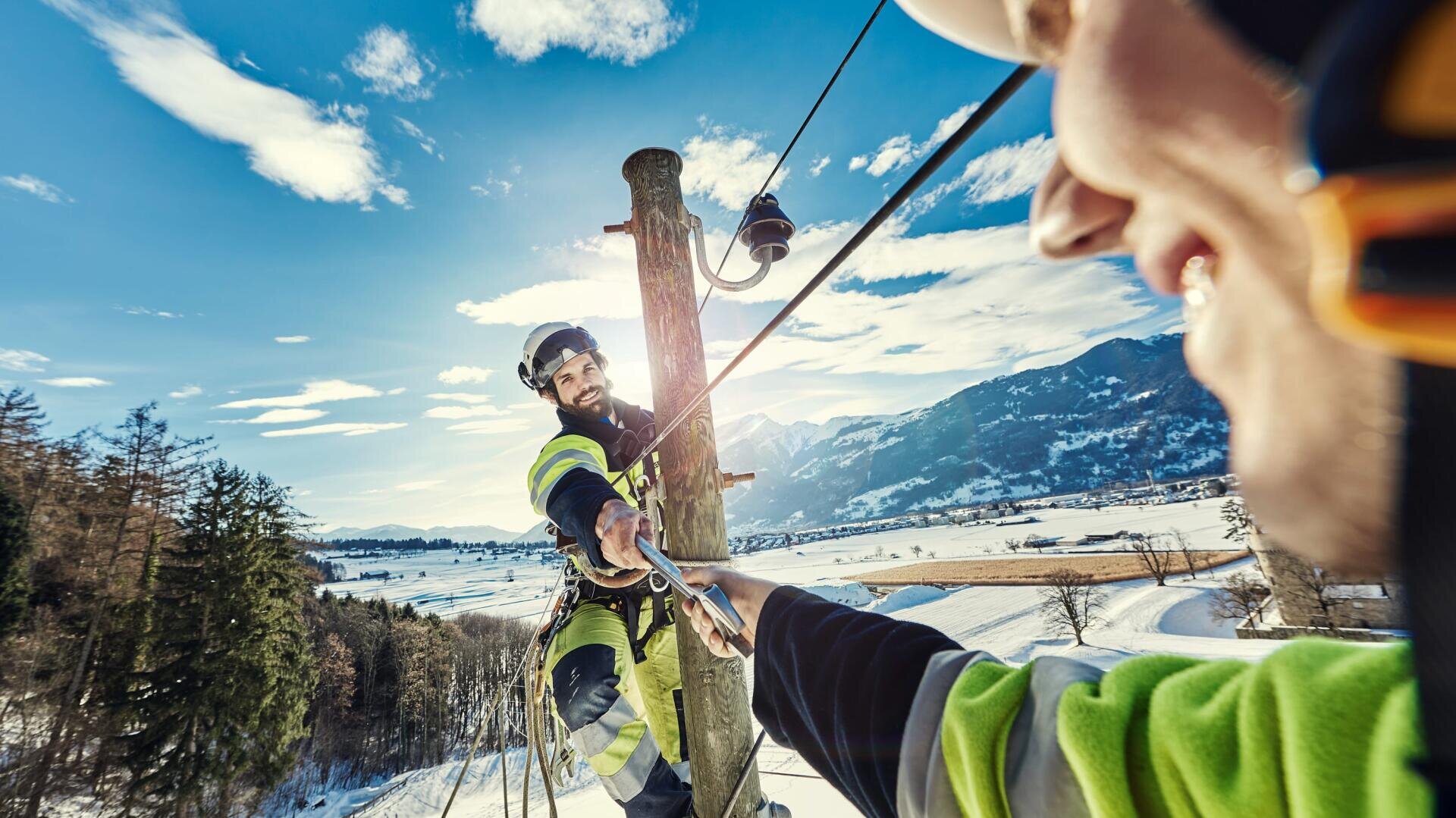 Zwei Arbeiter eines Versorgungsunternehmens, die Sicherheitsausrüstung tragen, arbeiten an einem Holzmast in einer verschneiten, bergigen Landschaft. Der eine Arbeiter reicht dem anderen ein Werkzeug oder ein Seil, während er mit Gurten unter einem hellen, sonnigen Himmel gesichert ist.