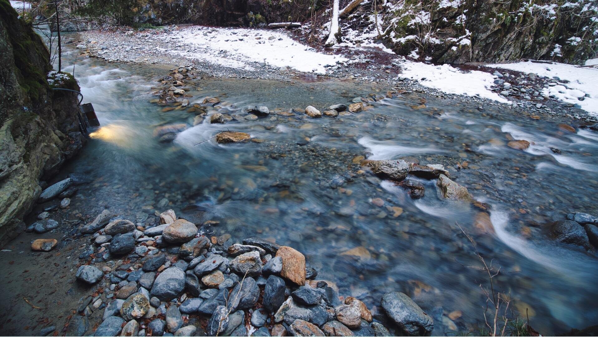 Ein klarer, felsiger Bach fließt durch eine winterliche Landschaft mit schneebedeckten Ufern und moosbewachsenen Felsen. Einige Steine bilden einen Teil des Weges durch das seichte Wasser, und ein sanftes Licht leuchtet von der linken Seite in der Nähe des Wasserrandes.