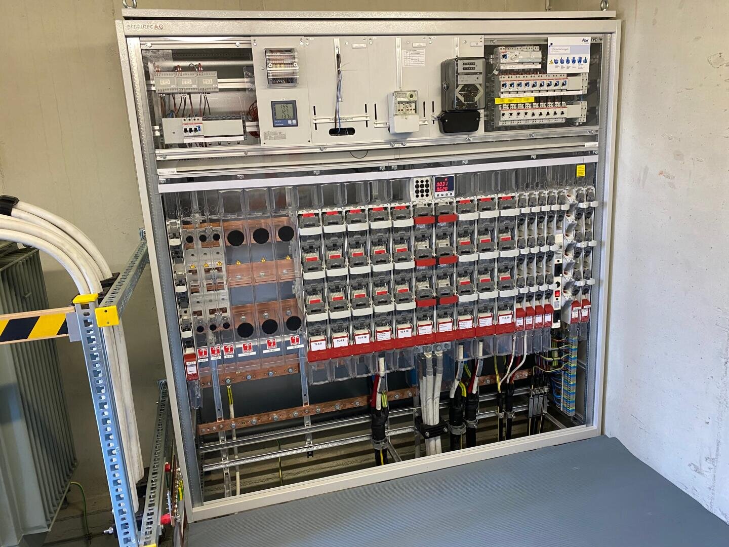 Eine offene elektrische Schalttafel mit mehreren Schaltern, Leistungsschaltern, Messgeräten und beschrifteten Kabeln, die in einem Metallrahmen an einer weißen Wand angebracht sind.