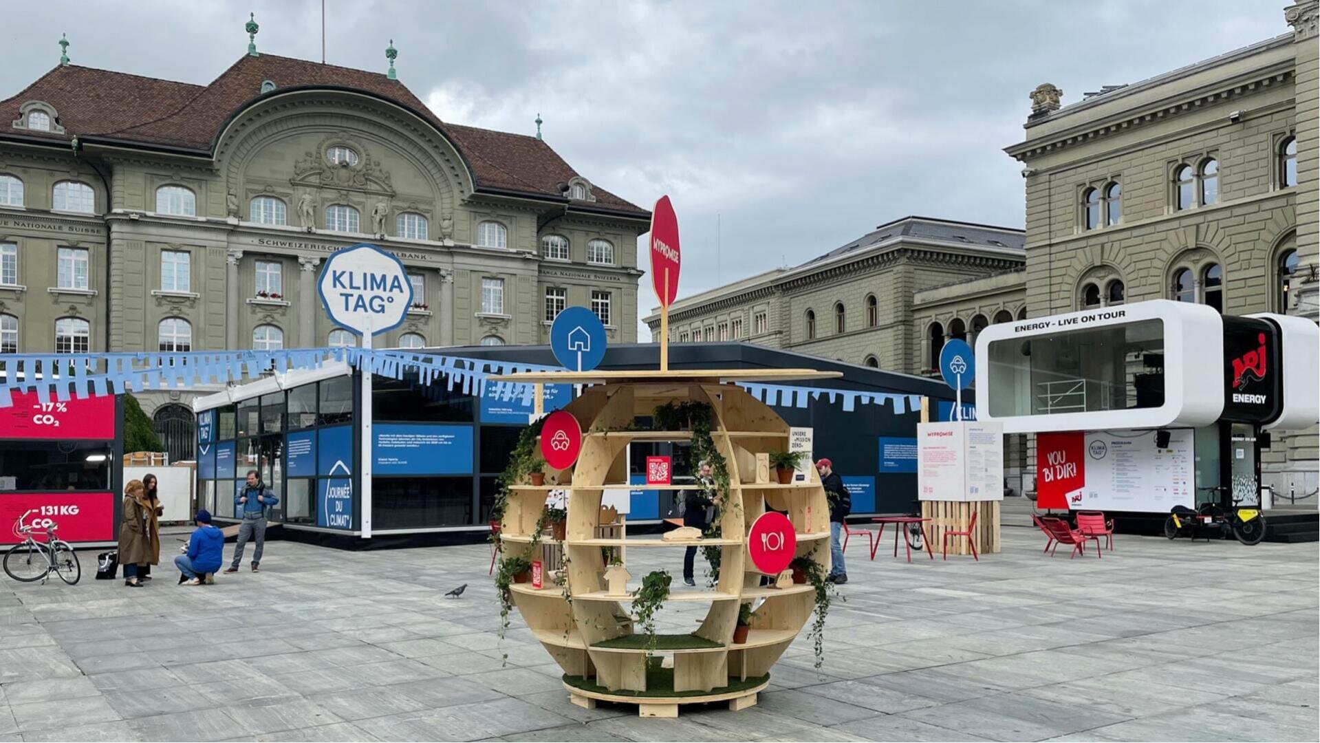 Ein öffentlicher Platz mit Exponaten zum Thema Klima, darunter eine runde Holzausstellung mit Pflanzen, Informationsständen, Bannern mit der Aufschrift KLIMA TAG und historischen Gebäuden im Hintergrund. Die Menschen gehen umher und interagieren mit den Ausstellungsstücken.