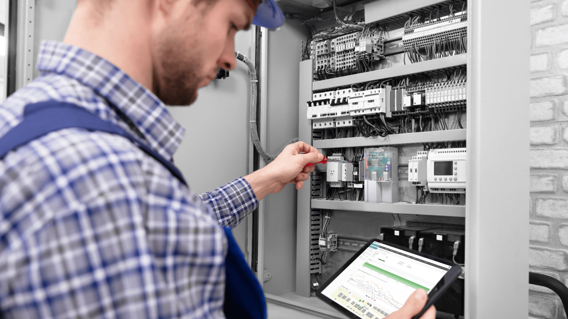 Ein Techniker im karierten Hemd arbeitet mit einem roten Schraubenzieher an einem geöffneten elektrischen Schaltschrank, während er ein Tablet hält, auf dem ein Diagramm angezeigt wird, das die Wartung oder Inspektion anzeigt.