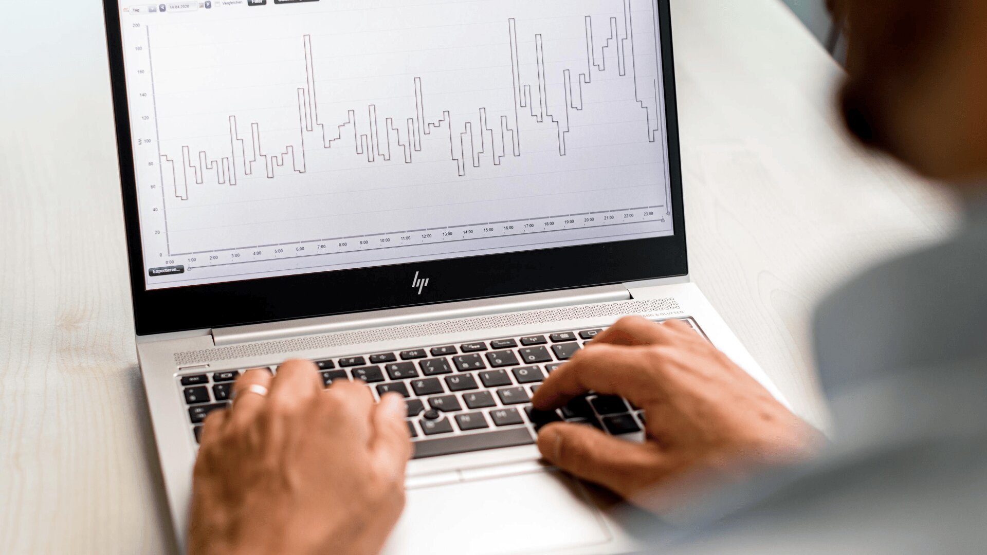 Eine Person tippt auf einem HP-Laptop, der ein Liniendiagramm mit wechselnden Spitzen und Tälern auf dem Bildschirm anzeigt, was auf Datenanalyse oder finanzielle Aktivitäten schließen lässt. Der Fokus liegt auf den Händen und der Laptop-Tastatur.