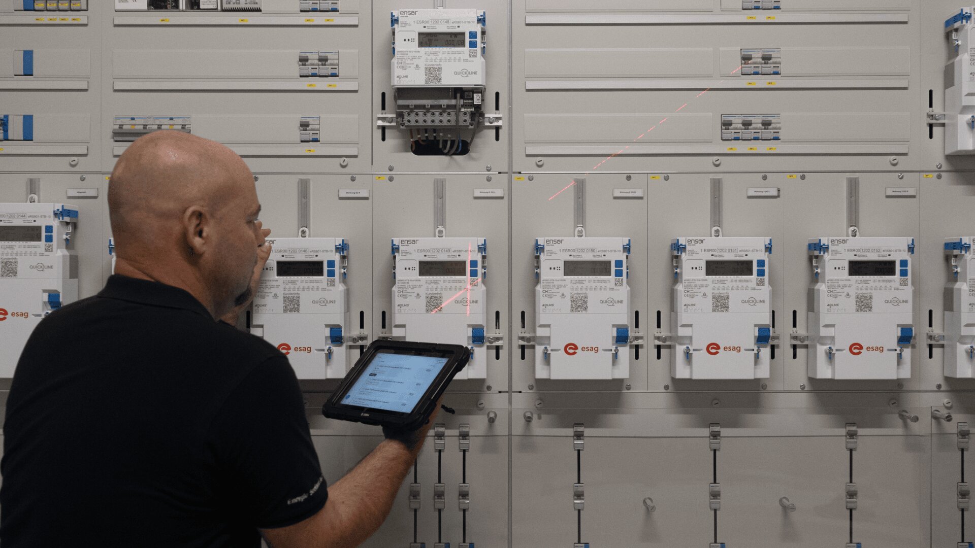 Eine Person verwendet ein Tablet und richtet einen roten Laser auf eine Reihe von digitalen Stromzählern, die auf einer Schalttafel montiert sind.