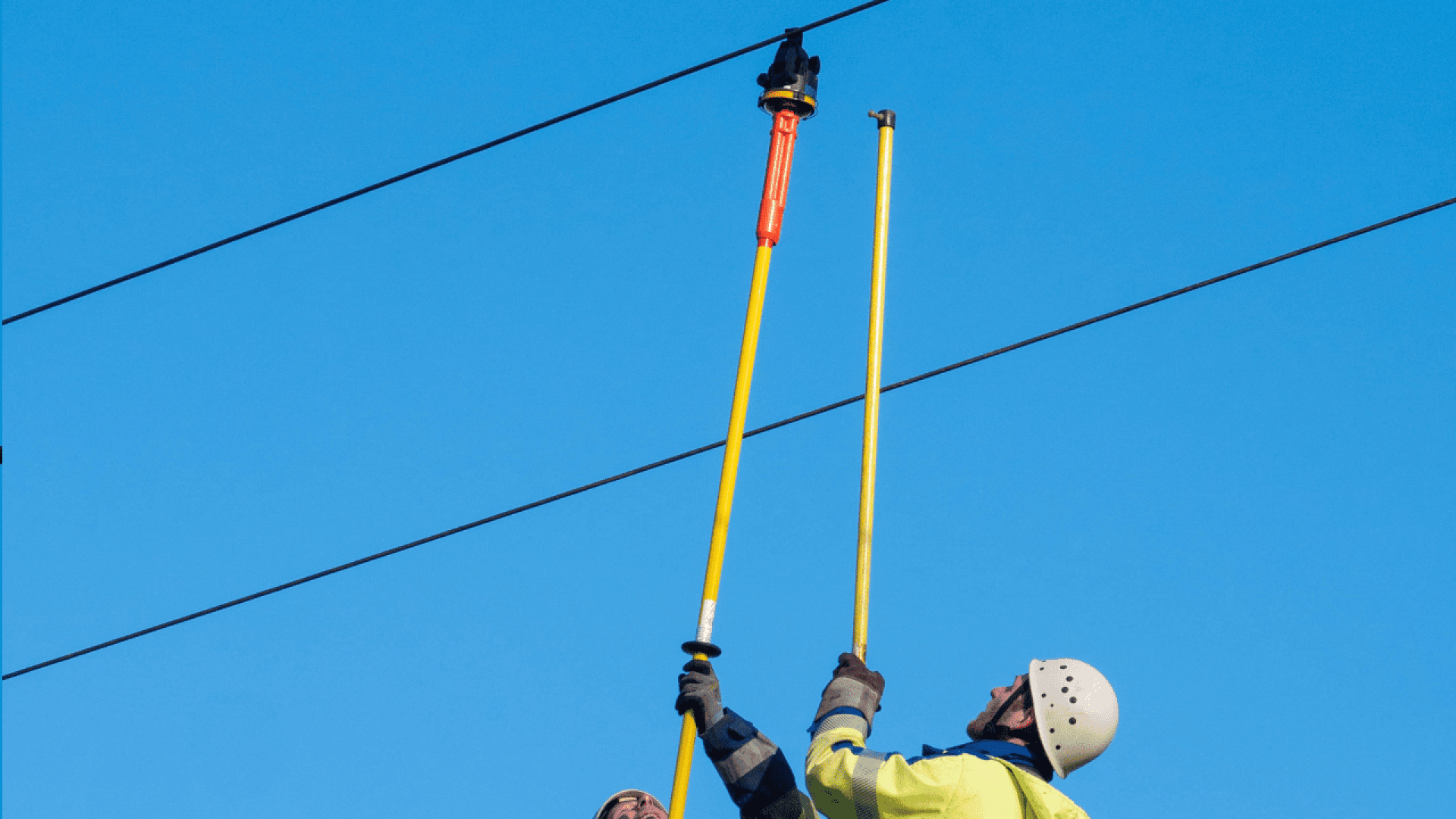 Zwei Arbeiter eines Energieversorgers in Schutzkleidung arbeiten mit isolierten Masten an Freileitungen bei strahlend blauem Himmel.