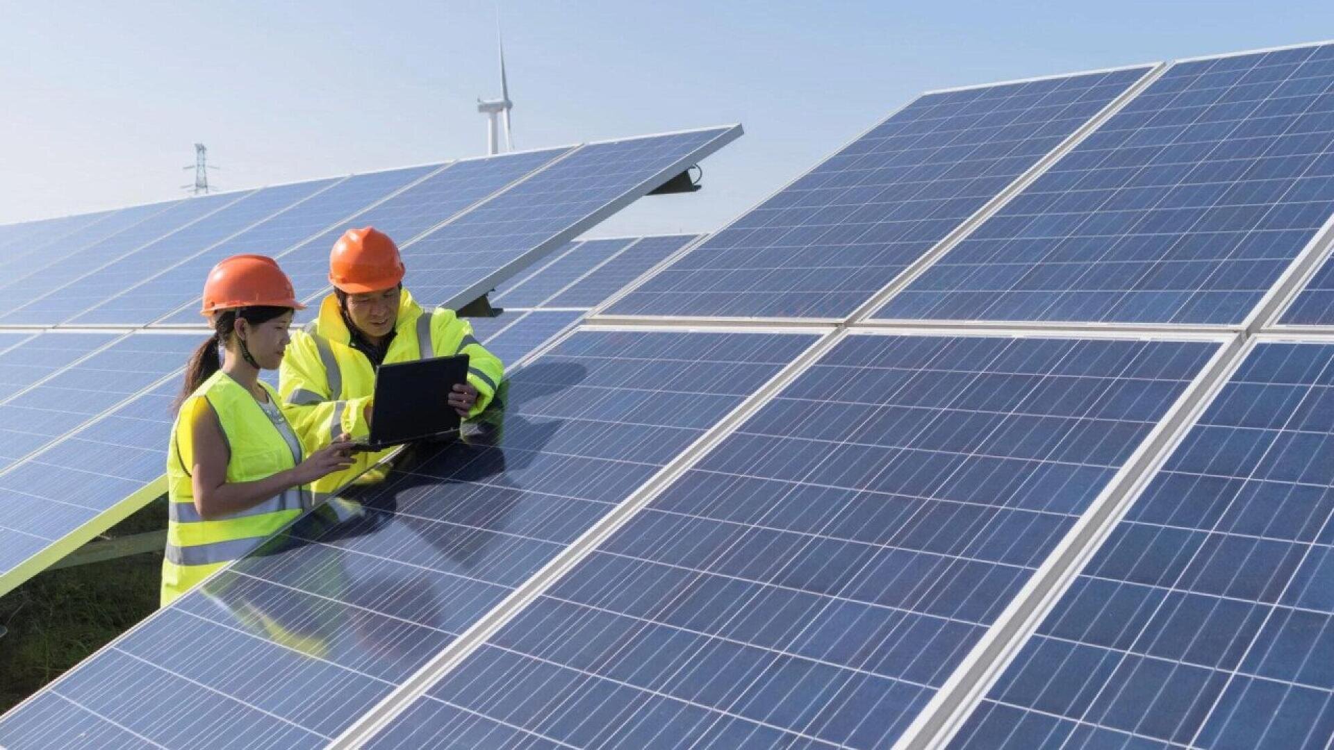 Two workers in safety vests and helmets examine a laptop while standing among large solar panels at a solar power facility, with a wind turbine visible in the background under a clear sky.