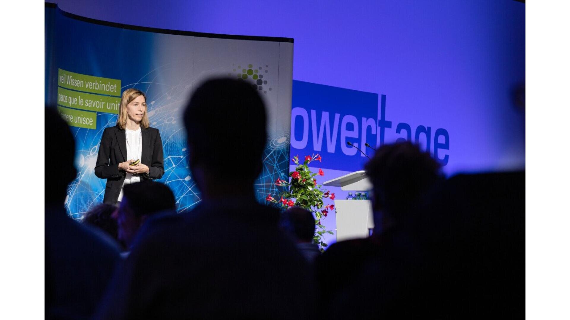 A woman in a suit stands on stage giving a presentation to an audience. The background features a blue screen with the word powertage and some plants and graphics. The audience is shown in silhouette.