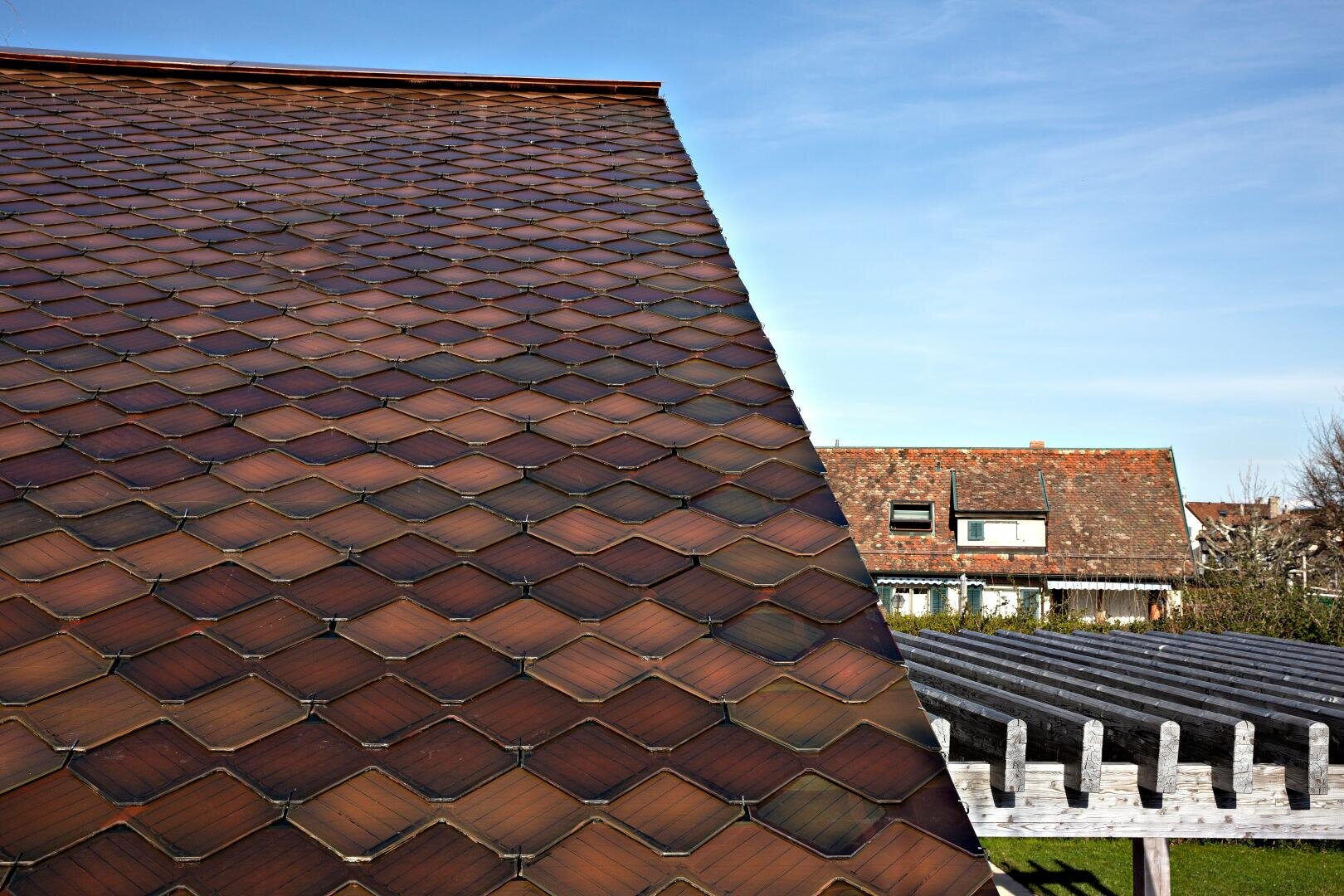 A close-up view of a sloped roof covered with brown fish scale tiles; in the background, another house with a similarly tiled roof and a wooden structure are visible under a clear blue sky.