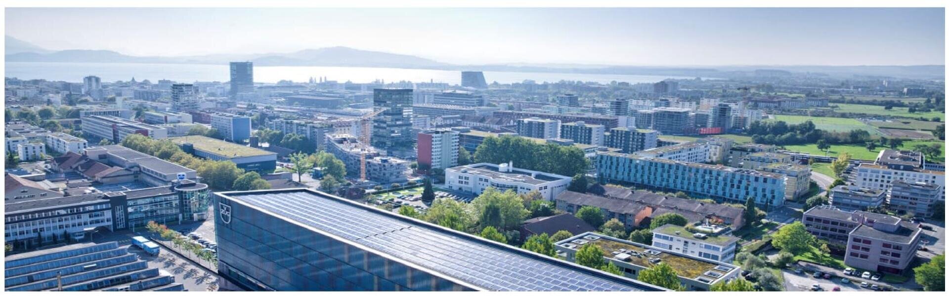Ein Panoramablick auf eine moderne Stadt mit zahlreichen Gebäuden, Grünflächen und entfernten Bergen an einem großen See unter klarem Himmel.