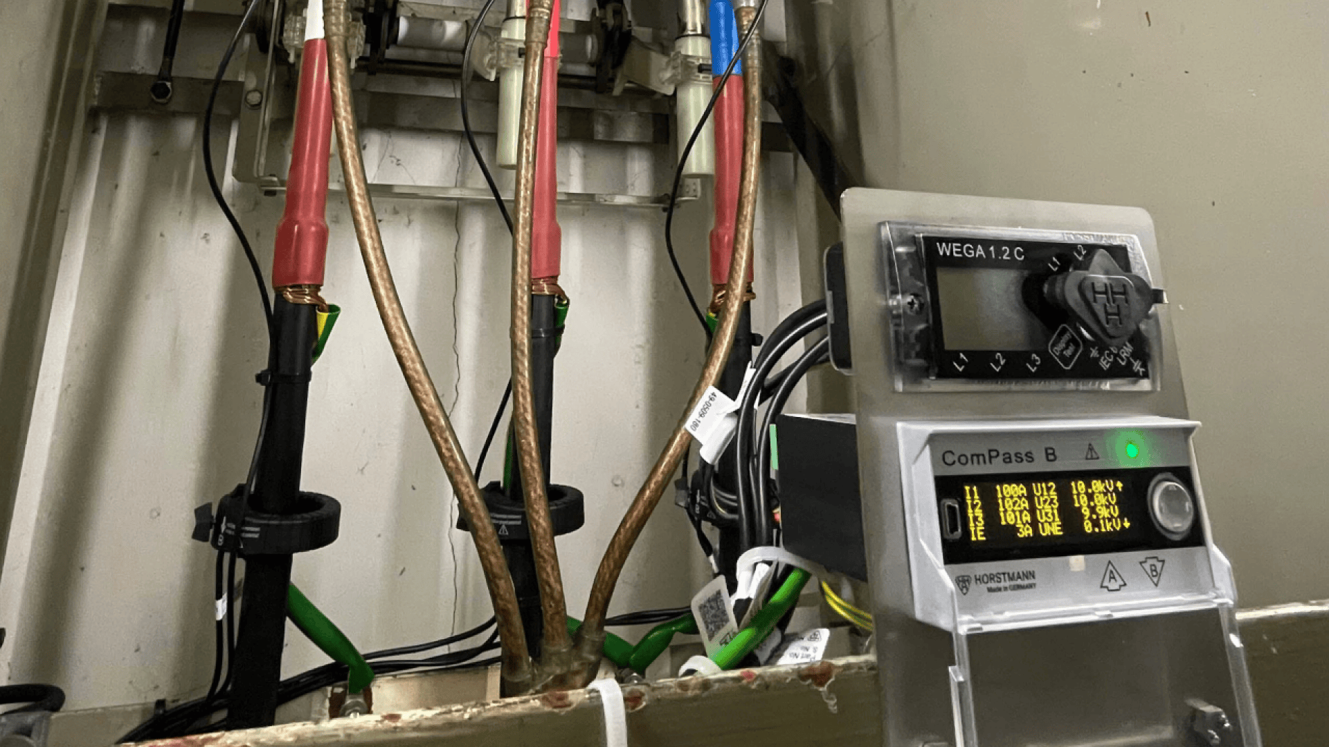 Eine Nahaufnahme von elektrischen Geräten, darunter Kabel mit roter Isolierung, ein digitales Messgerät, das die Werte anzeigt, und verschiedene grüne und schwarze Kabel, die mit Geräten in einem Schaltschrank verbunden sind.