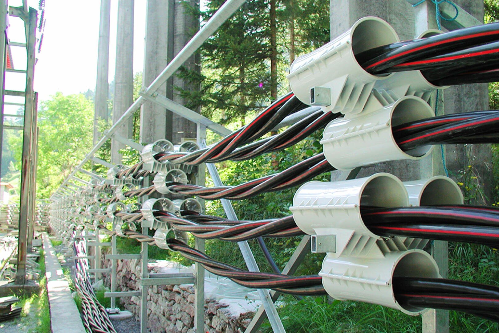 Multiple bundles of large black electrical cables are neatly organized and held in place by white plastic cable clamps on an outdoor metal framework, with greenery and concrete pillars visible in the background.
