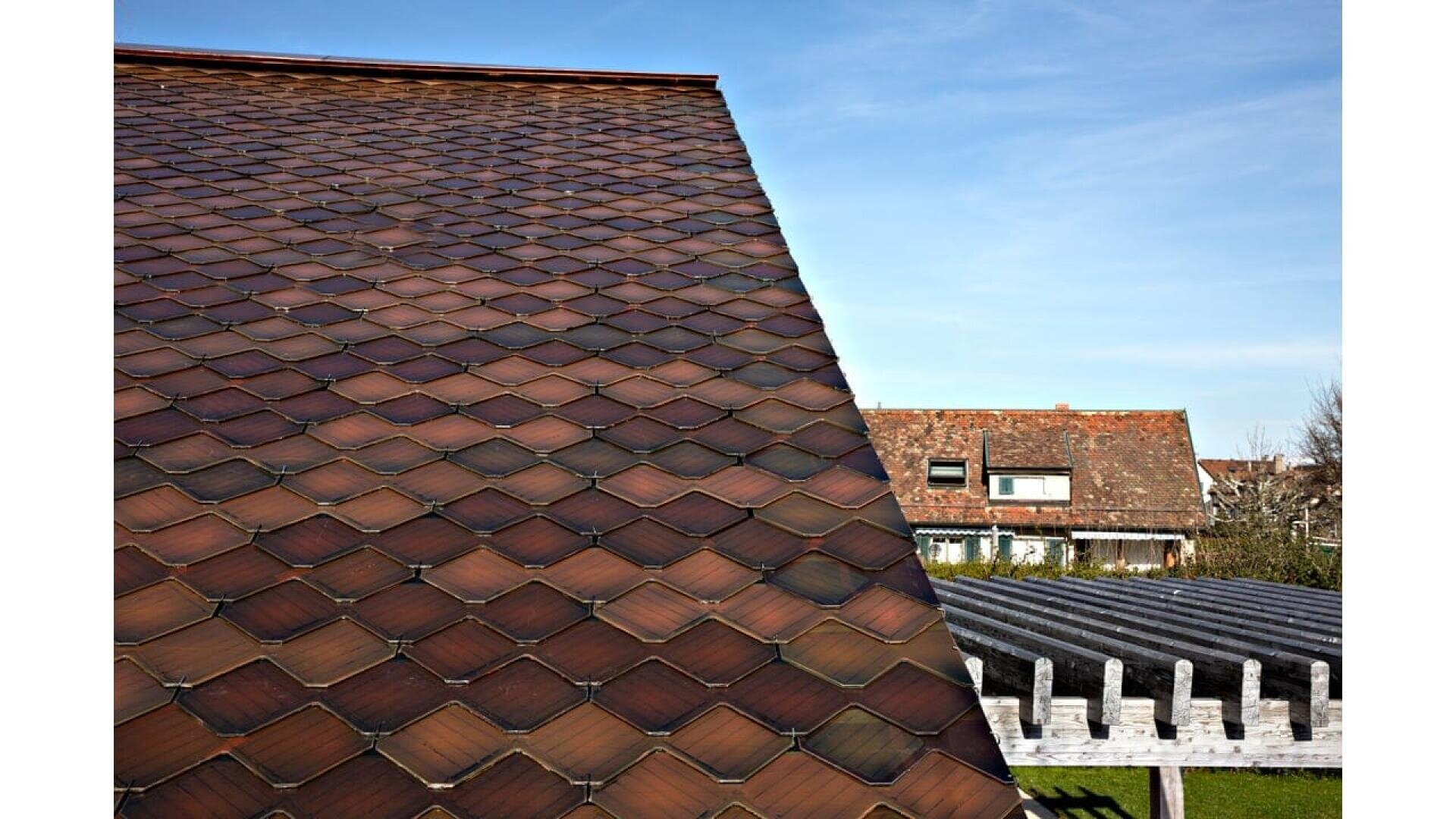 Nahaufnahme eines steil abfallenden Daches mit dunklen, rautenförmigen Ziegeln; im Hintergrund sind Häuser und ein blauer Himmel zu sehen.
