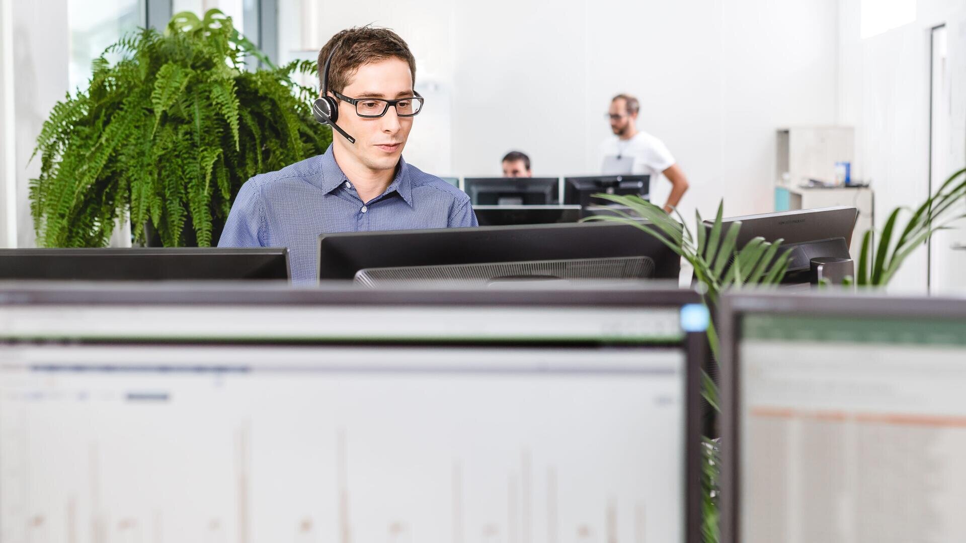 Ein Mann mit Brille und Headset arbeitet an einem Computer in einem hellen Büro mit mehreren Monitoren, großen Grünpflanzen und anderen Personen, die im Hintergrund arbeiten.