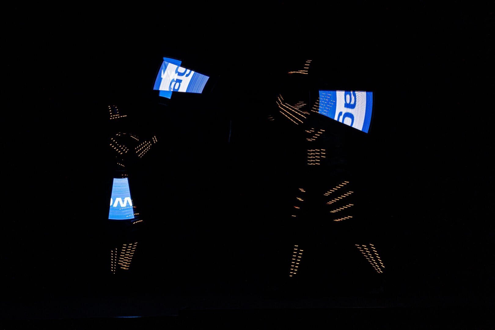 Zwei Performer in der Dunkelheit tragen Anzüge mit leuchtenden orangefarbenen Lichtern und halten beleuchtete blaue und weiße LED-Paneele, die abstrakte, futuristische Formen vor einem schwarzen Hintergrund erzeugen.