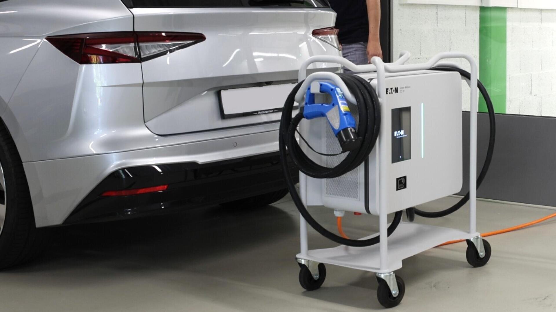A portable electric vehicle charging station on wheels is positioned near the rear of a silver car in an indoor setting. The charger has a screen, cables, and a plug ready for charging.