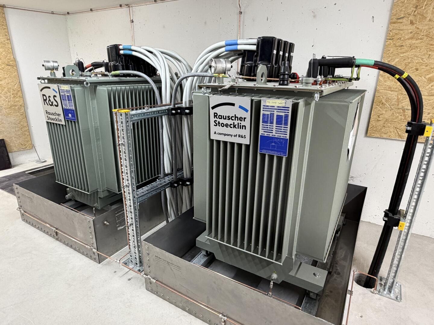 Zwei große elektrische Industrietransformatoren mit der Aufschrift "Rauscher & Stoecklin" sind in einem Innenraum installiert, mit dicken Kabeln verbunden und auf Metallplattformen vor einer weißen, holzgetäfelten Wand montiert.
