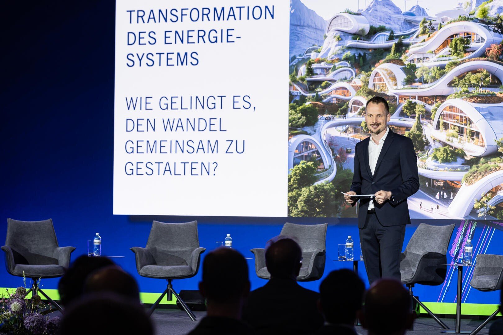 Ein Mann steht auf der Bühne und hält Papiere neben vier leeren Stühlen in der Hand. Hinter ihm ist ein Bildschirm mit einem deutschen Text über die Energiewende zu sehen. Auf der Leinwand ist auch eine futuristische Stadt abgebildet. Vorne sitzen die Zuhörer.