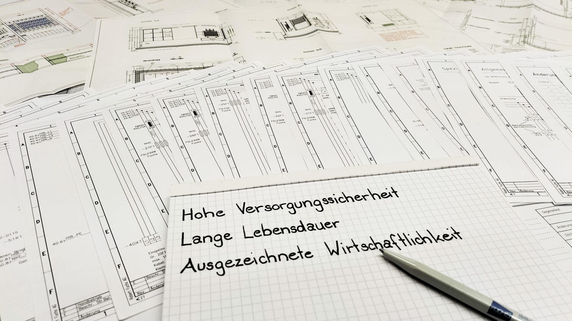 Ein Stapel technischer Zeichnungen liegt auf einem Tisch. Im Vordergrund ist ein Rasterpapier mit deutschem handgeschriebenem Text zu sehen: Hohe Versorgungssicherheit, Lange Lebensdauer, Ausgezeichnete Wirtschaftlichkeit. In der Nähe ruht ein Stift.