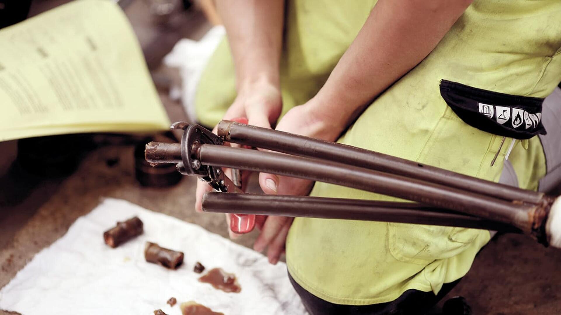 Eine Person in einem gelben Hemd kniet auf dem Boden und repariert mit Werkzeugen ein Metallstativ oder ein Geräteteil. In der Nähe liegen verschiedene Kleinteile und ein Stück weißes Tuch ausgebreitet. Im Hintergrund ist ein gelbes Papier mit Text zu sehen.
