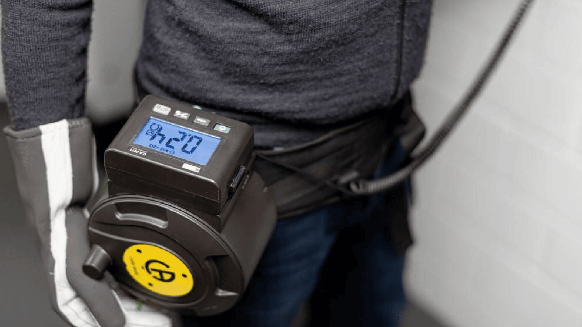 Eine Person mit Schutzhandschuhen hält einen Geigerzähler mit einer Digitalanzeige von 2,70, die die Strahlungswerte anzeigt. Das Gerät ist mit einem Gurt an der Taille der Person befestigt.