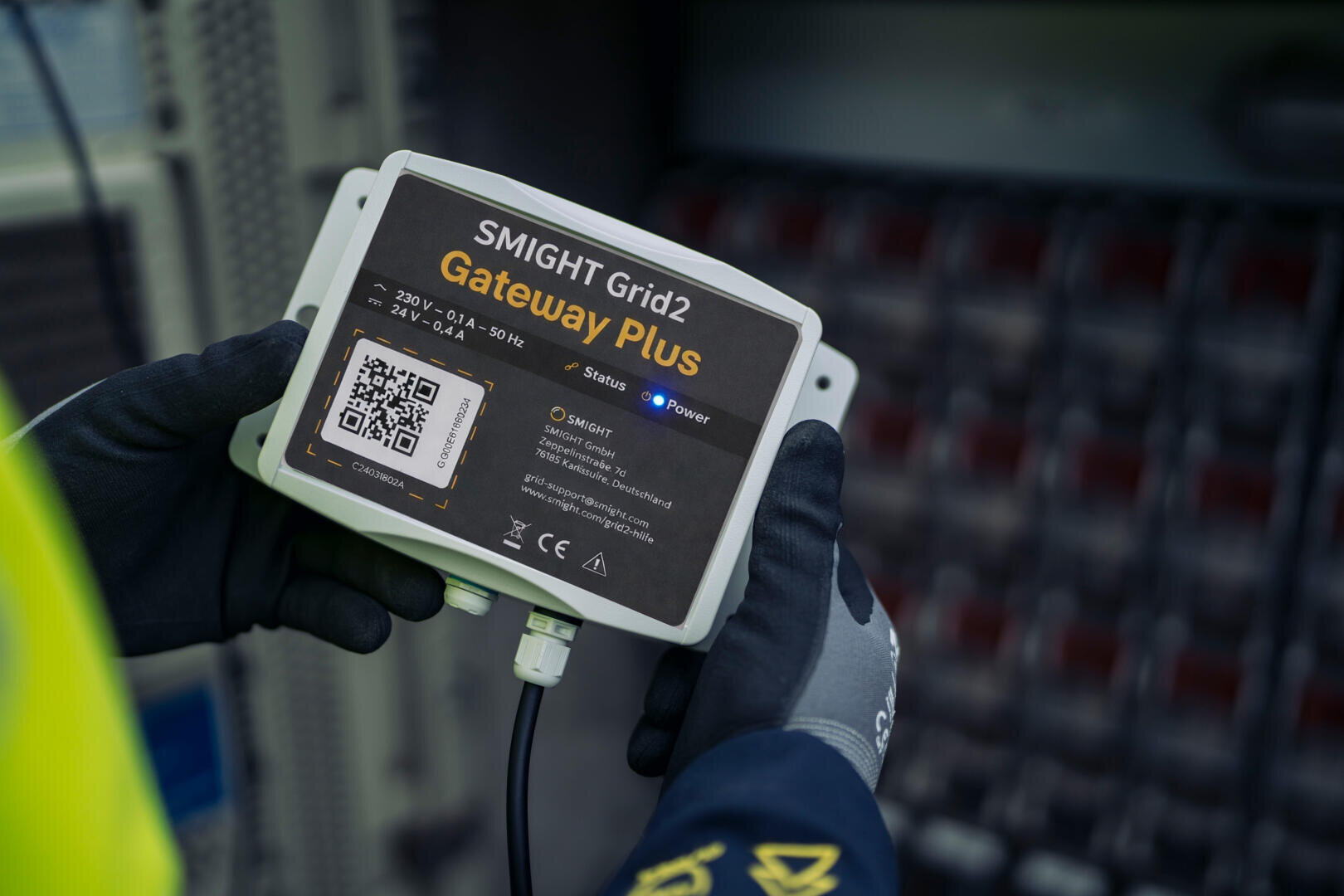 A person wearing gloves holds a SMIGHT Grid2 Gateway Plus device, which has indicator lights and a QR code on the label, in front of an electrical device.