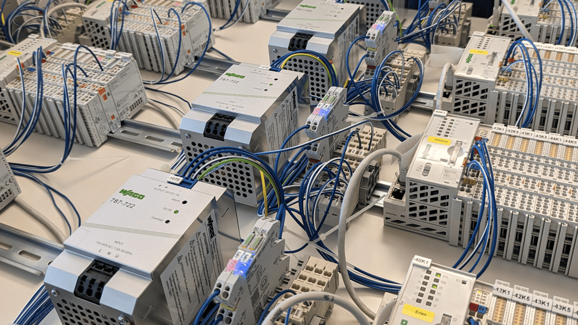 Nahaufnahme eines industriellen Schaltschranks mit mehreren miteinander verbundenen elektronischen Modulen, Verdrahtungsklemmen und blauen Kabeln, die auf Metallschienen angeordnet sind und eine komplexe Automatisierungs- oder Elektroanlage zeigen.