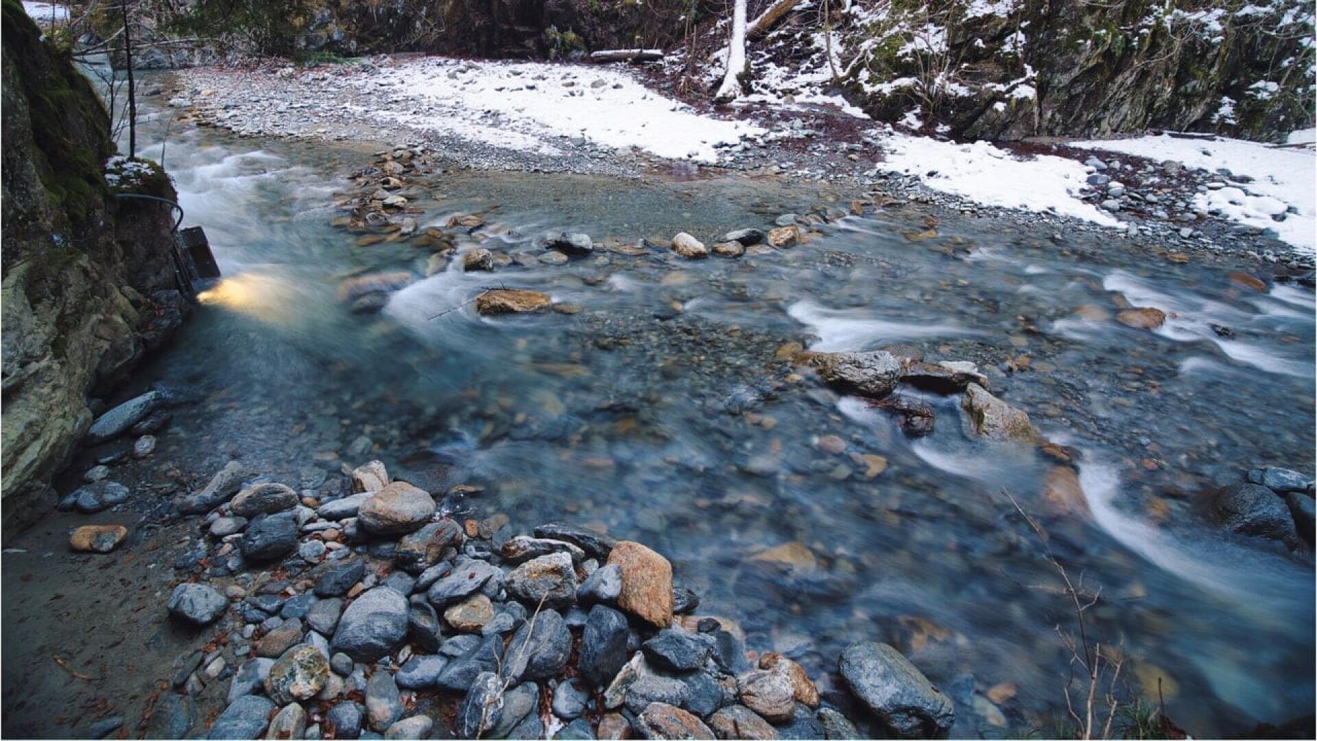 A shallow, rocky stream flows through a forested area with snow on the banks and ground. Water rushes around large stones, while wintery trees and moss-covered rocks line the background.