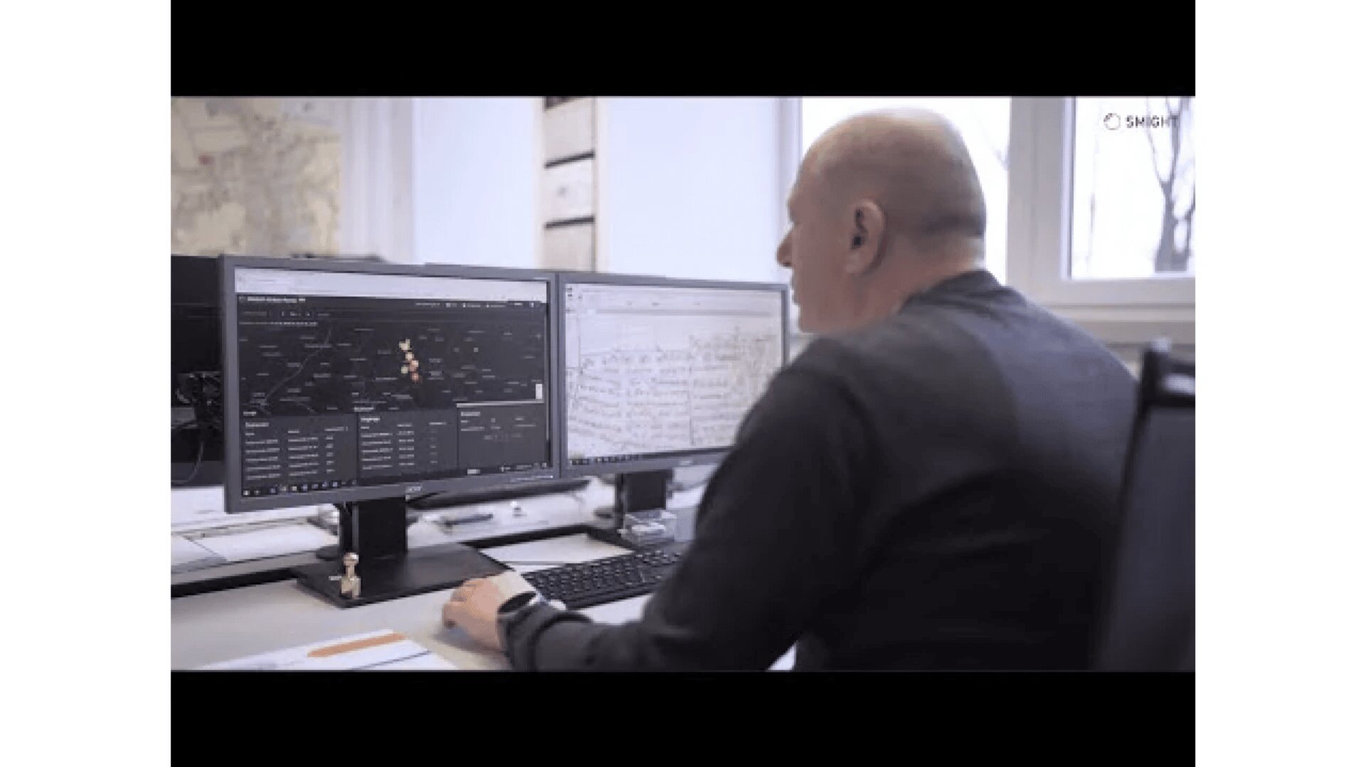 Ein Mann sitzt an einem Schreibtisch und arbeitet an einem Computer mit zwei Monitoren. Ein Bildschirm zeigt eine dunkle Oberfläche mit Diagrammen oder Daten, während der andere ein Dokument mit handschriftlichen Notizen anzeigt. Auf dem Schreibtisch liegen Büromaterialien.