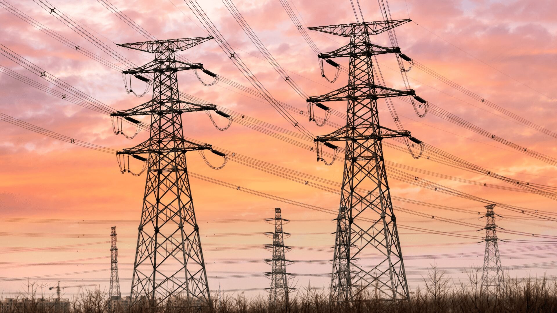 Hochspannungsmasten und -leitungen erstrecken sich bei Sonnenuntergang über die Landschaft, mit einem farbenfrohen Himmel in Orange-, Pink- und Violetttönen im Hintergrund.