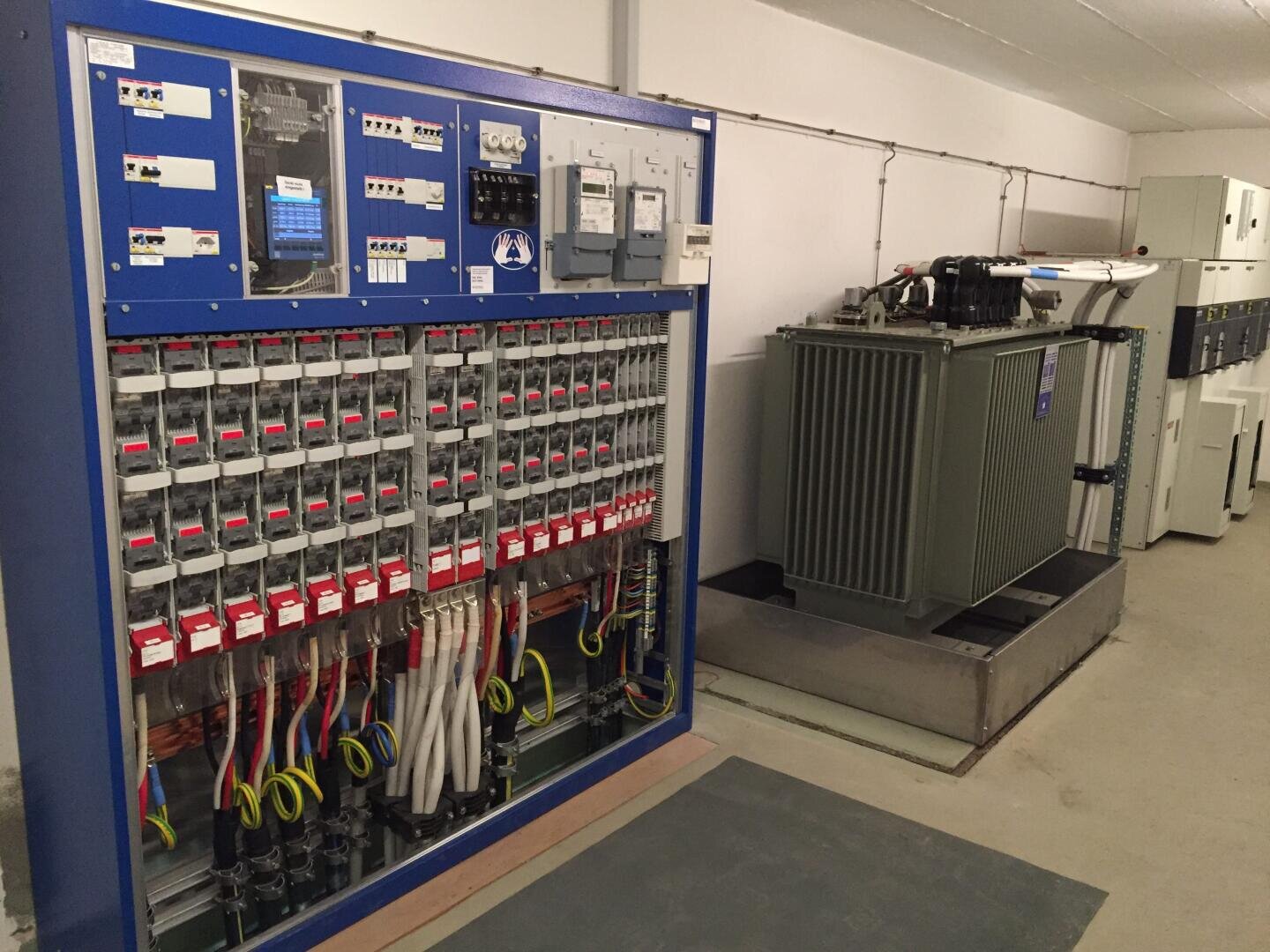 Elektrische Schalttafel mit mehreren Schaltern und beschrifteten Kabeln auf der linken Seite und ein großer Industrietransformator auf der rechten Seite, beide in einem sauberen, gut beleuchteten Raum mit weißen Wänden und einem gefliesten Boden installiert.