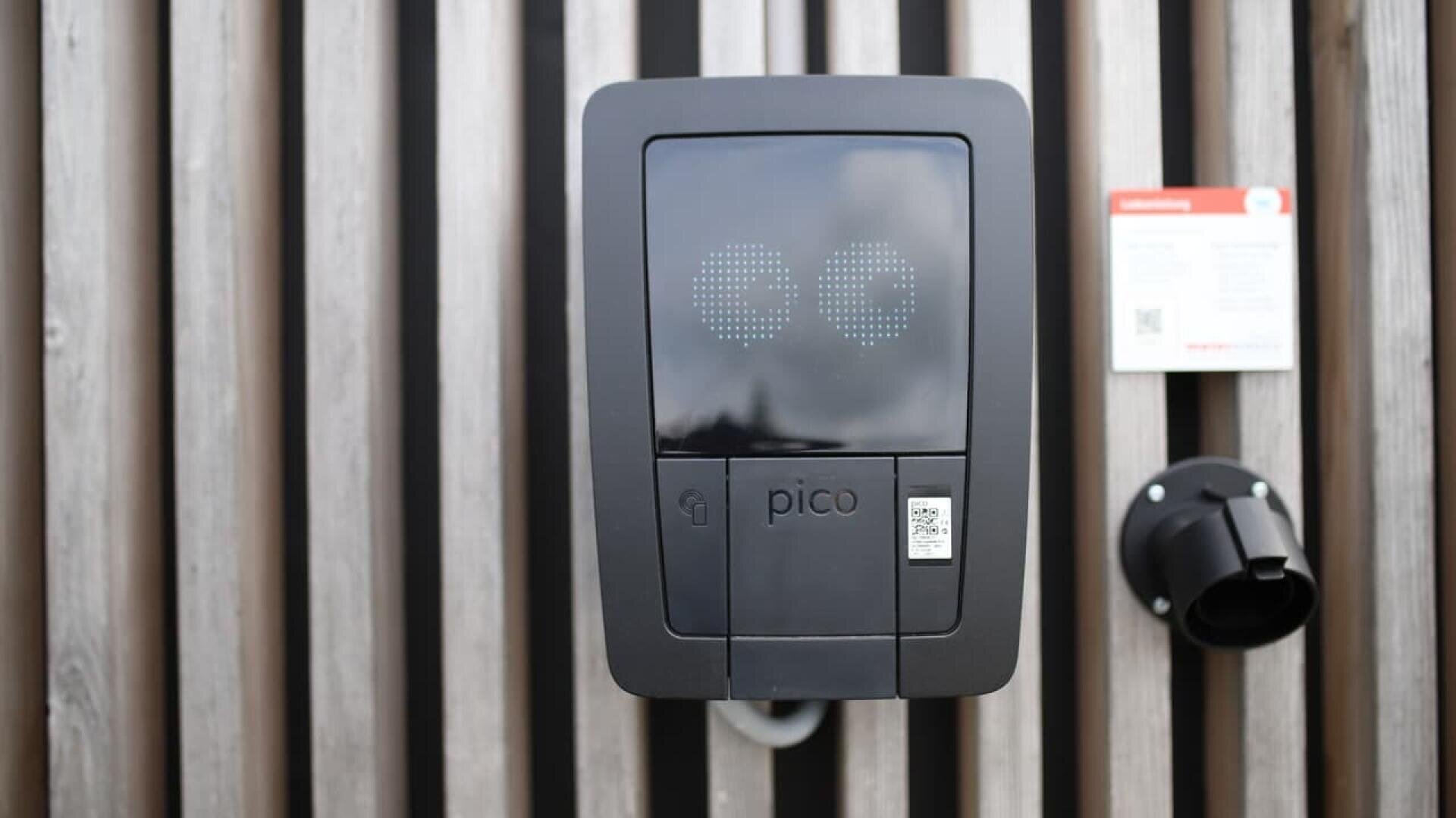A black Pico electric vehicle charging station is mounted on a wooden slat wall, with a round socket on the right and a small instruction sign posted above it.