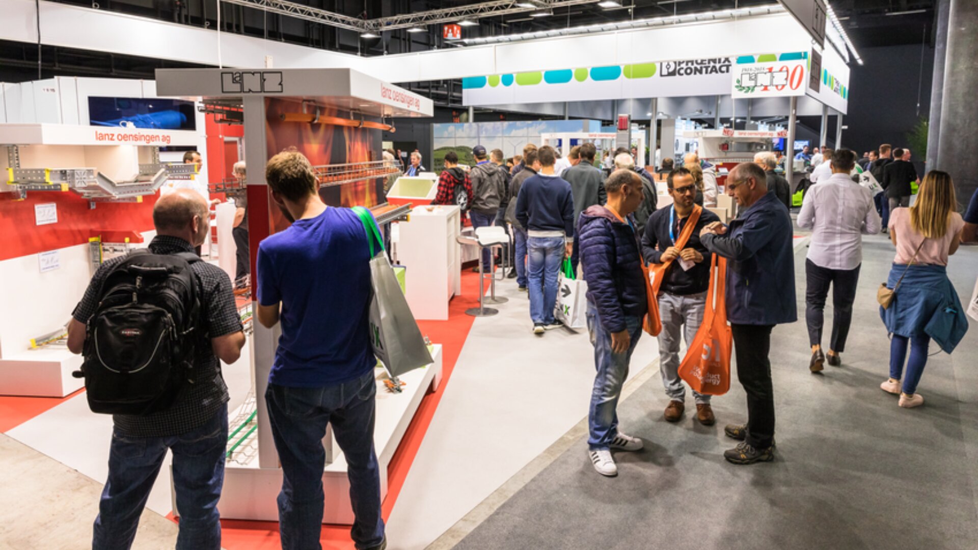 Eine Gruppe von Menschen geht auf einer belebten Messe oder Ausstellung umher und tauscht sich aus, umgeben von Ständen, Produktauslagen und Bannern bei heller Beleuchtung. Einige tragen orangefarbene Tragetaschen und sind leger gekleidet.