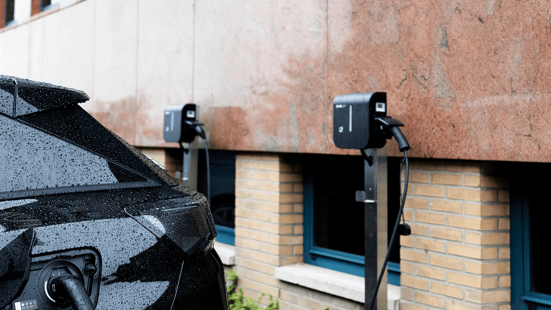 Ein schwarzes Auto wird an einer Ladestation für Elektrofahrzeuge aufgeladen, die an einer Wand außerhalb eines Gebäudes mit Ziegel- und roten Steinwänden angebracht ist. Regentropfen sind auf dem Auto zu sehen. Im Hintergrund befindet sich eine weitere Ladestation.
