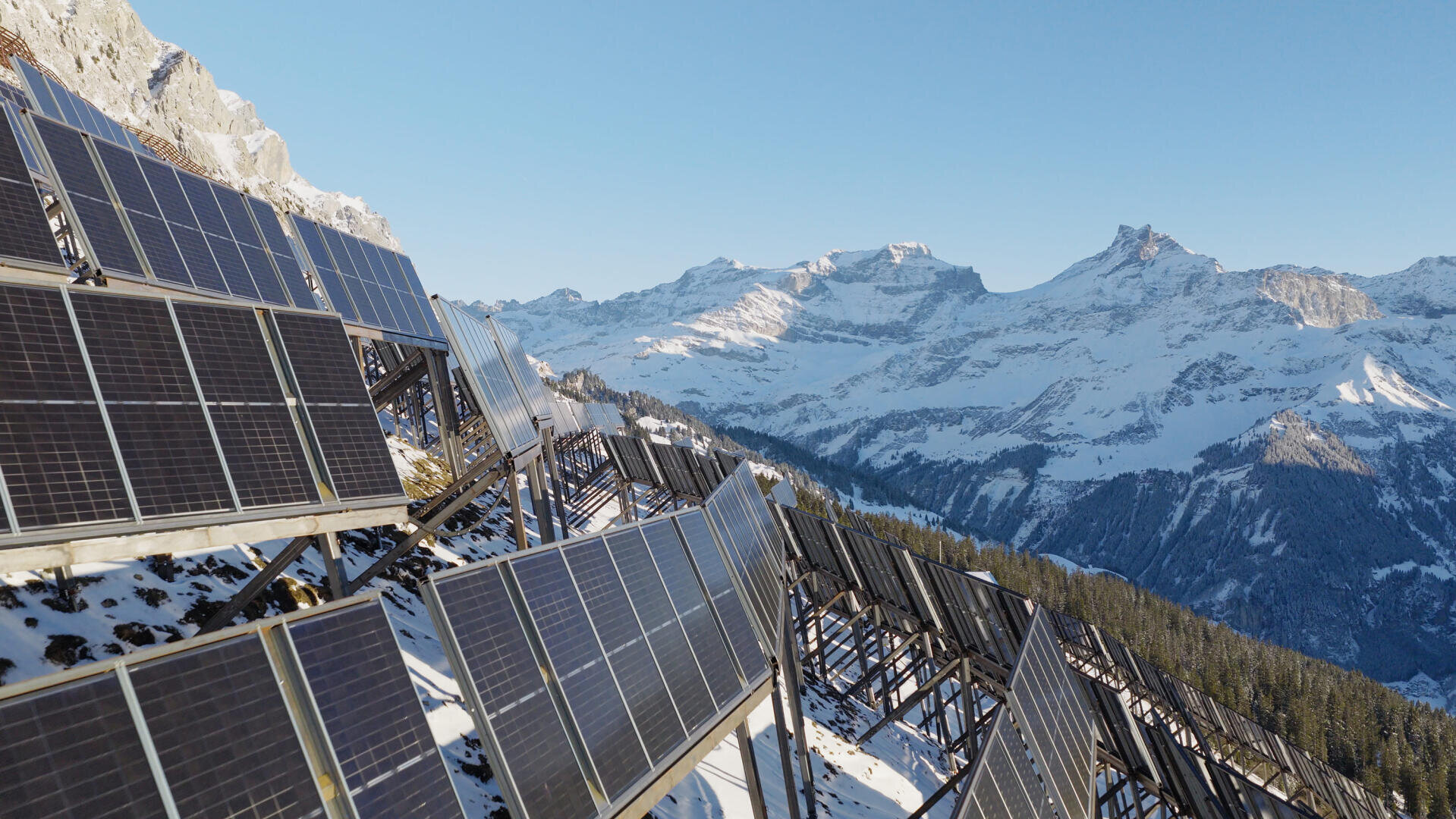 Auf einem schneebedeckten Berghang installierte Solarmodule mit hohen, schroffen, schneebedeckten Gipfeln im Hintergrund unter einem klaren blauen Himmel.