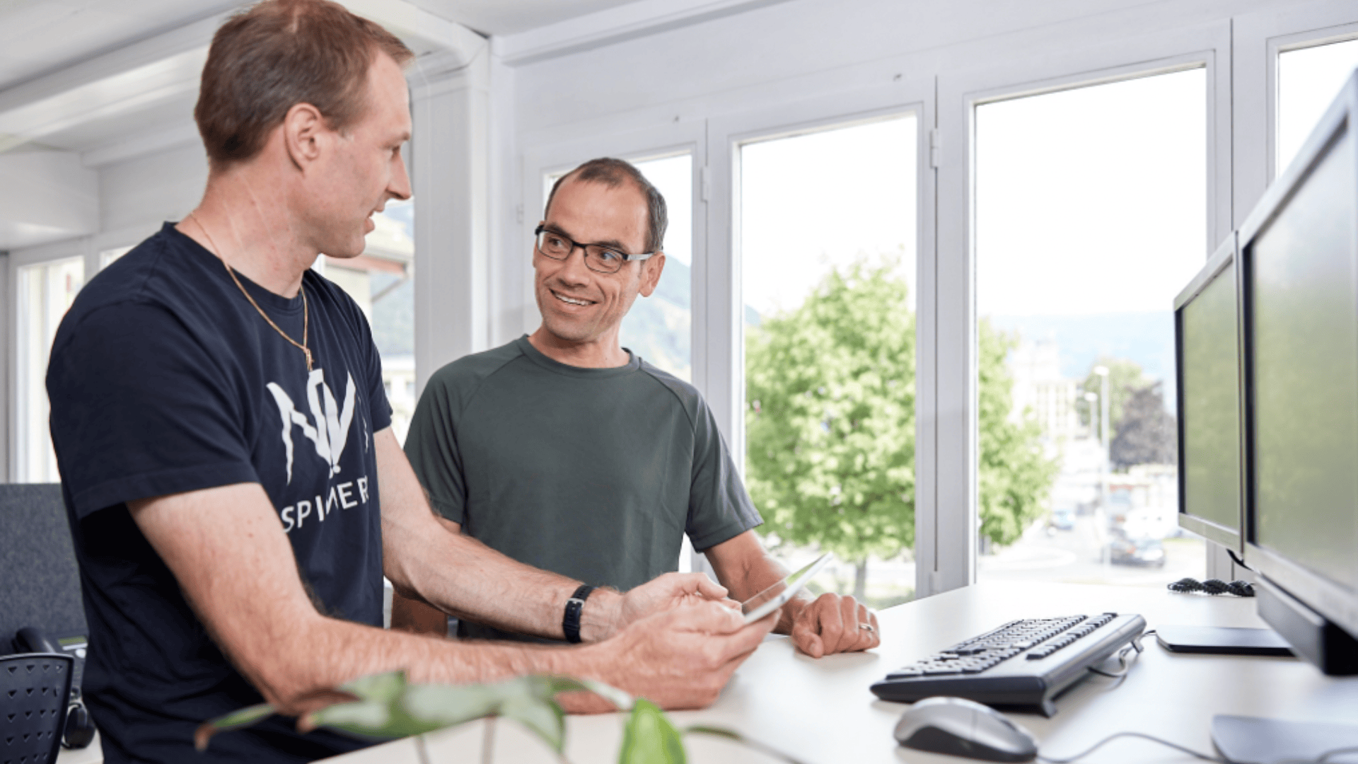 Zwei Männer unterhalten sich und lächeln in einem hellen Büroraum. Ein Mann hält ein Smartphone in der Hand, während der andere mit seinen Händen gestikuliert. Auf dem Schreibtisch vor ihnen befinden sich Computermonitore, eine Tastatur und eine Maus. Im Hintergrund sind große Fenster zu sehen.