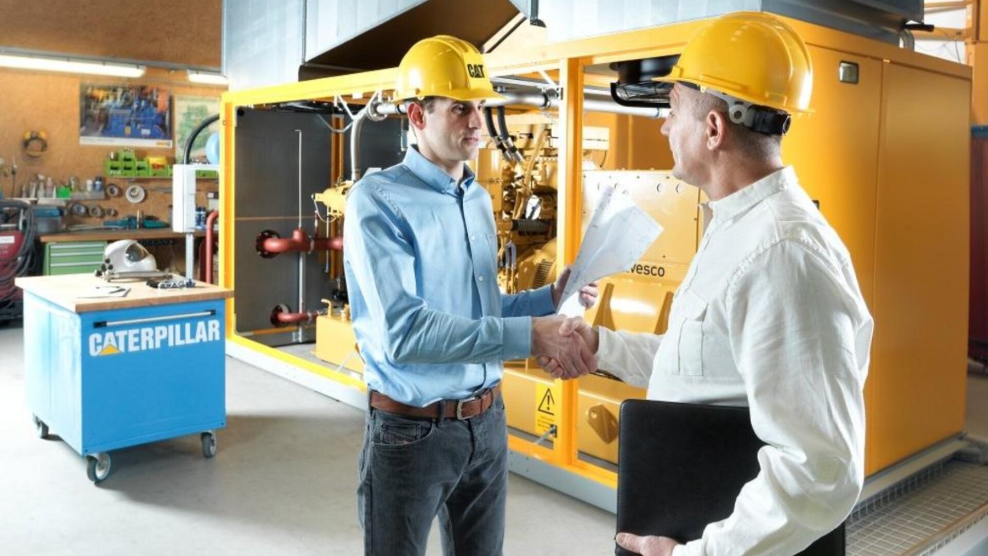 Zwei Männer mit gelben Schutzhelmen geben sich vor einer großen gelben Caterpillar-Maschine in einer Industriehalle die Hand. Der eine hält Dokumente in der Hand, der andere einen schwarzen Ordner.
