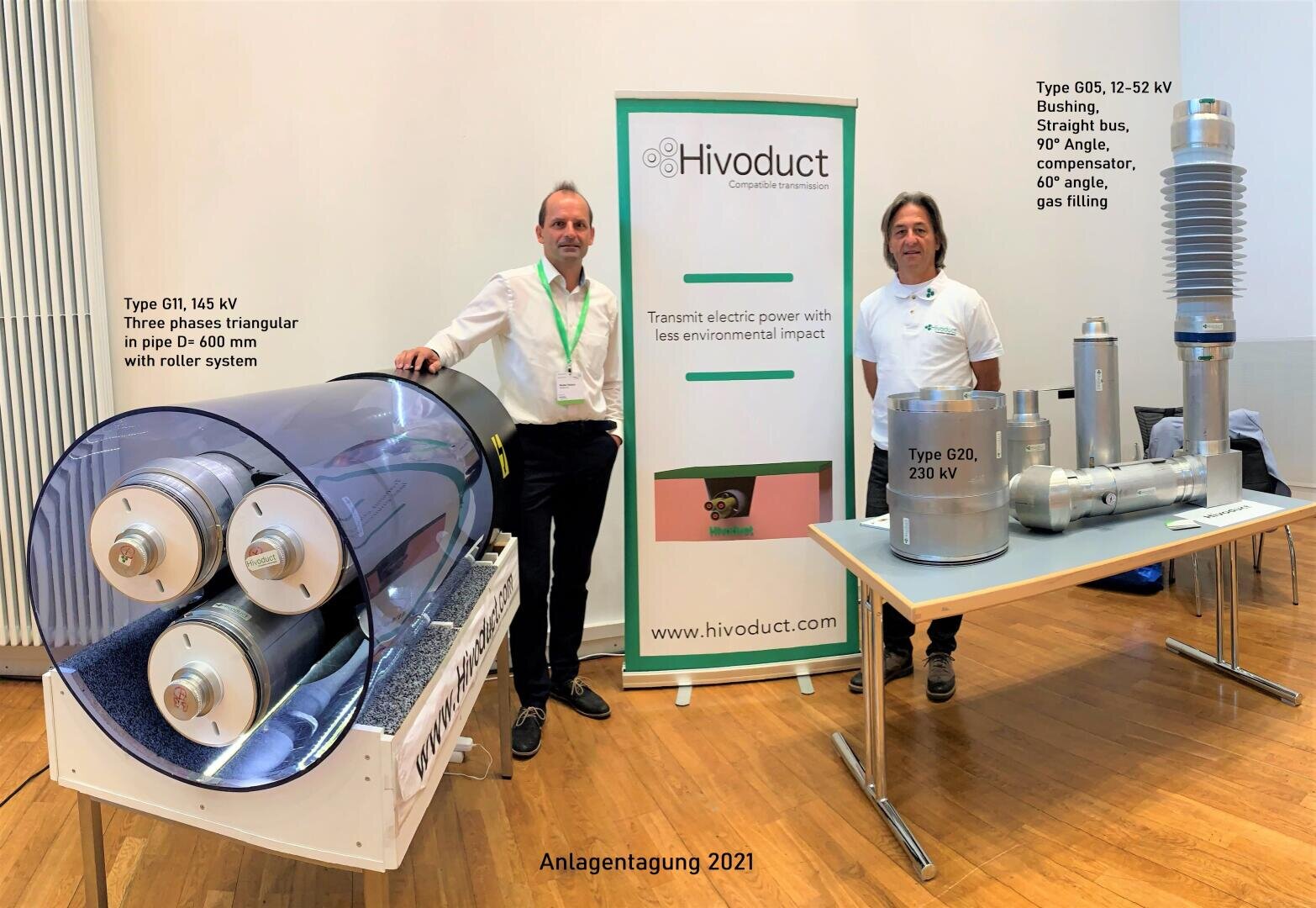 Zwei Männer stehen vor einer Ausstellung von Elektrogeräten und einem Banner von Hivoduct. Auf der linken Seite ist ein großes Kabelmodell zu sehen, auf der rechten Seite verschiedene Hochspannungskomponenten. Es scheint sich um eine Ausstellung oder Konferenz zu handeln.