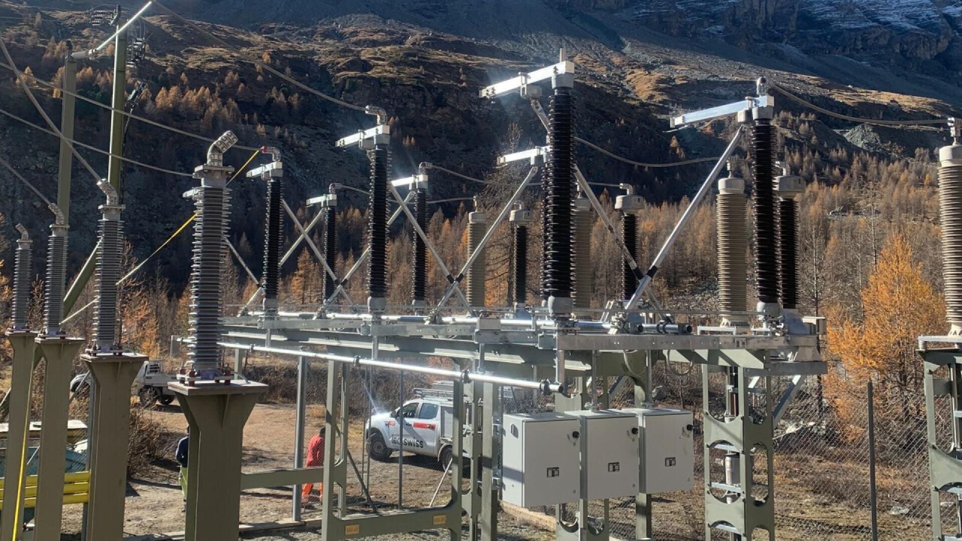 Electrical substation with metal structures, insulators, and control boxes set outdoors in a mountainous area, surrounded by autumn-colored trees and a parked white vehicle in the background.