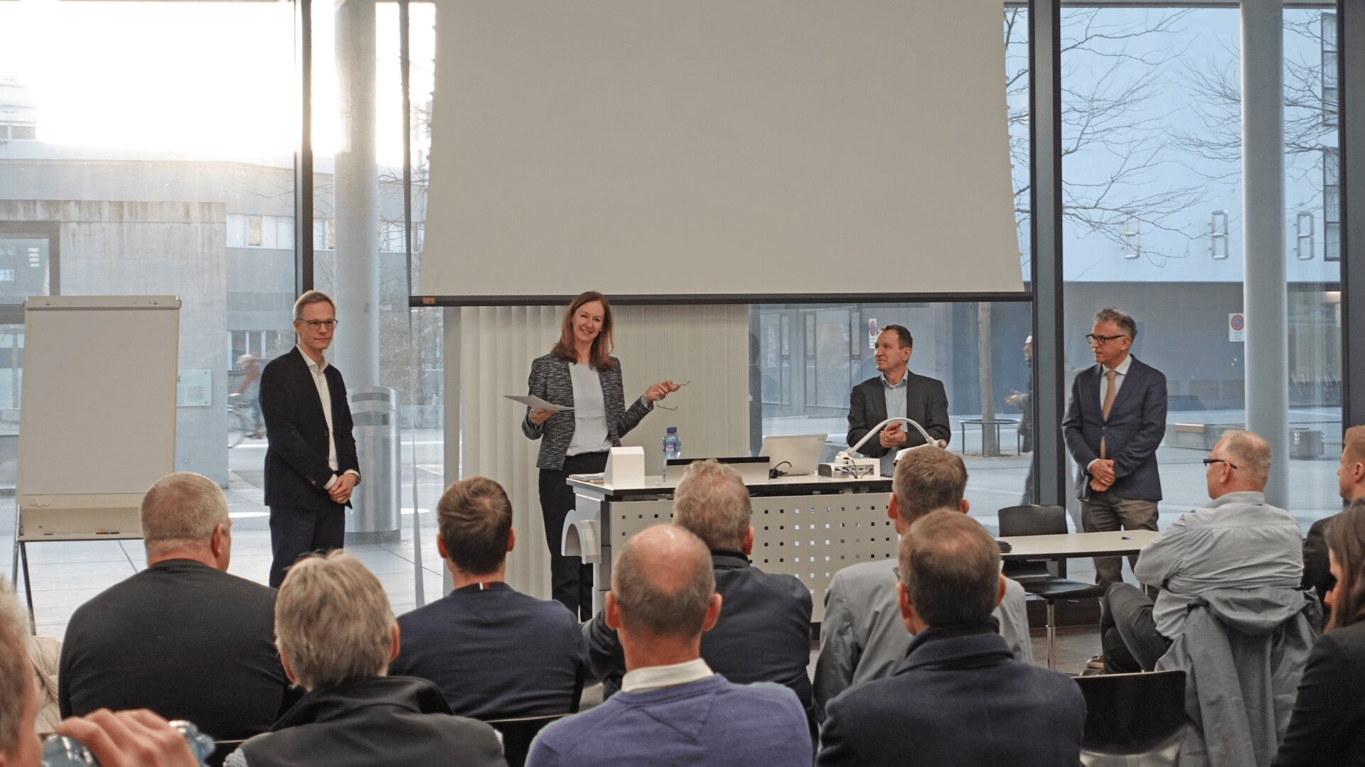 Eine Frau steht vorne in einem Raum und spricht zu einem Publikum, flankiert von drei Männern. Sie befinden sich in einem modernen Konferenzraum mit großen Fenstern und einem Präsentationsbildschirm hinter ihnen.
